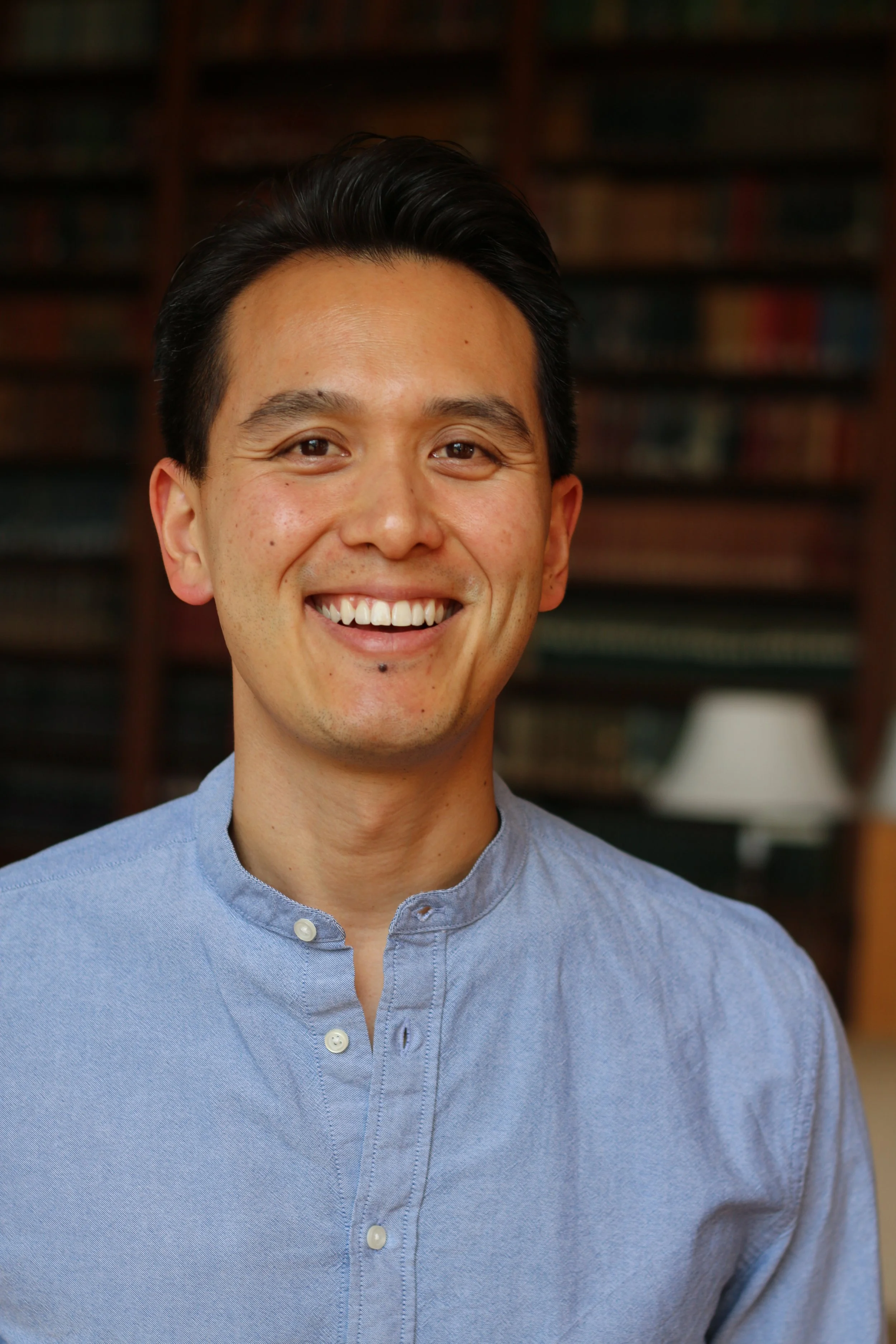 Close-up of a smiling Asian man in a light blue button-up shirt, standing indoors with a blurred background of bookshelves and a lamp.