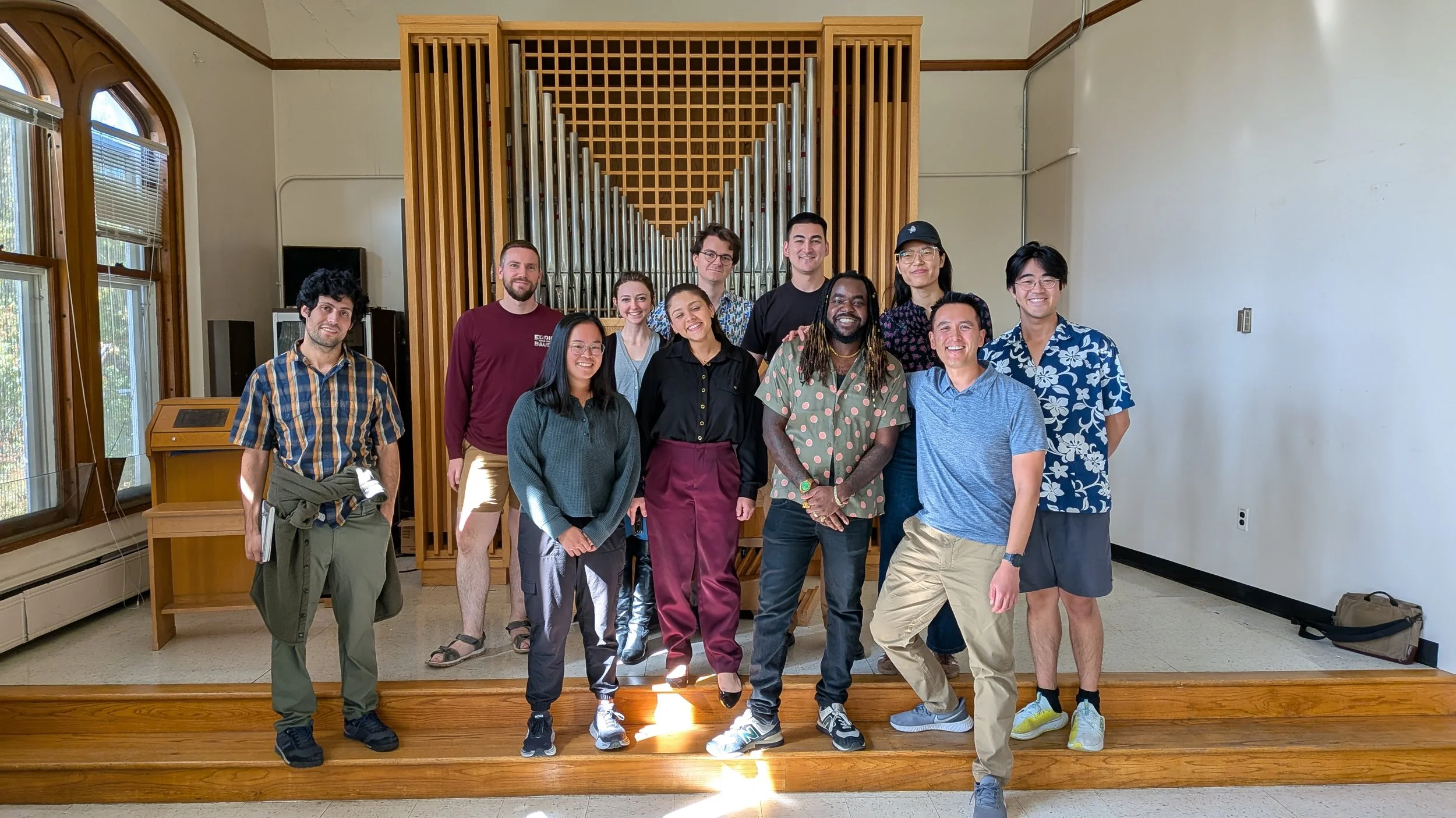 A diverse group of people standing together for a photo inside a room with a wooden organ behind them, windows to the left, and a plain white wall to the right.
