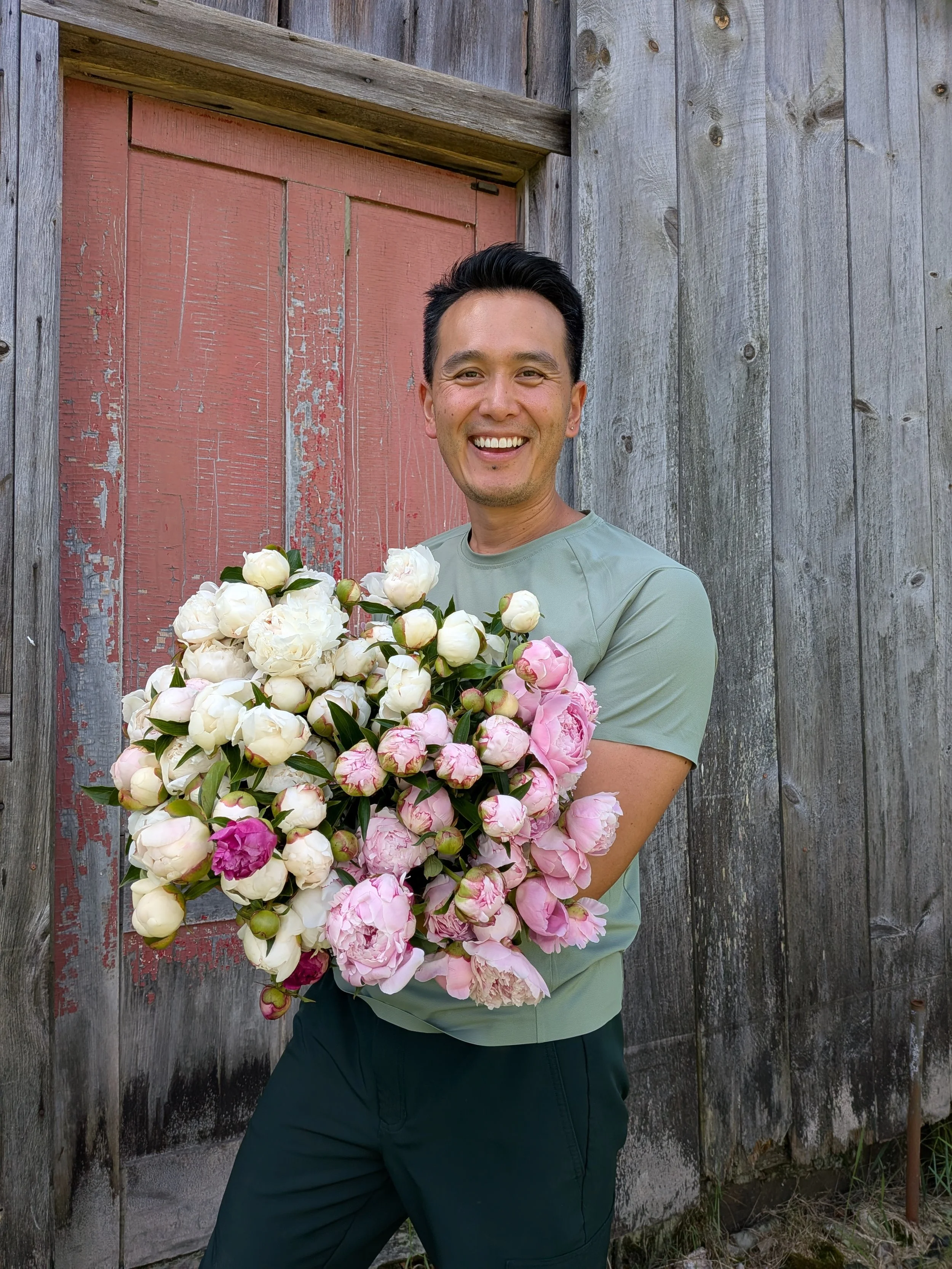 A man smiling and holding a large bouquet of white and pink peonies in front of a weathered wooden fence and red door.