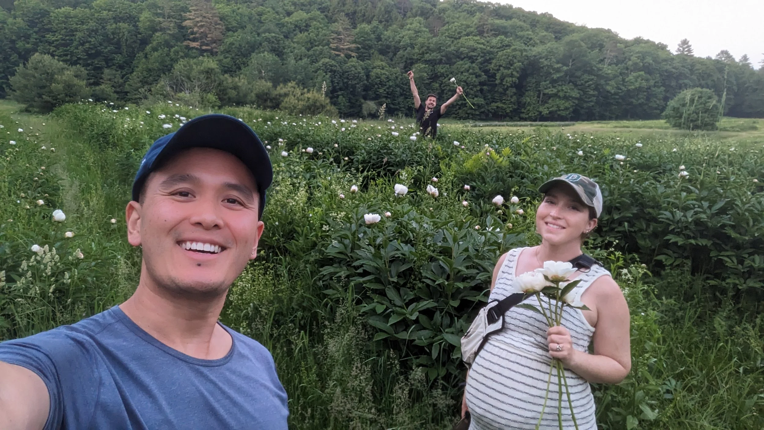 Three people in a lush garden with green trees and flowering bushes, smiling and taking a selfie. One young man in a blue shirt and cap is in the foreground. A young woman with a hat and striped top is holding flowers, and a man in the background appears to be celebrating with his arms raised.