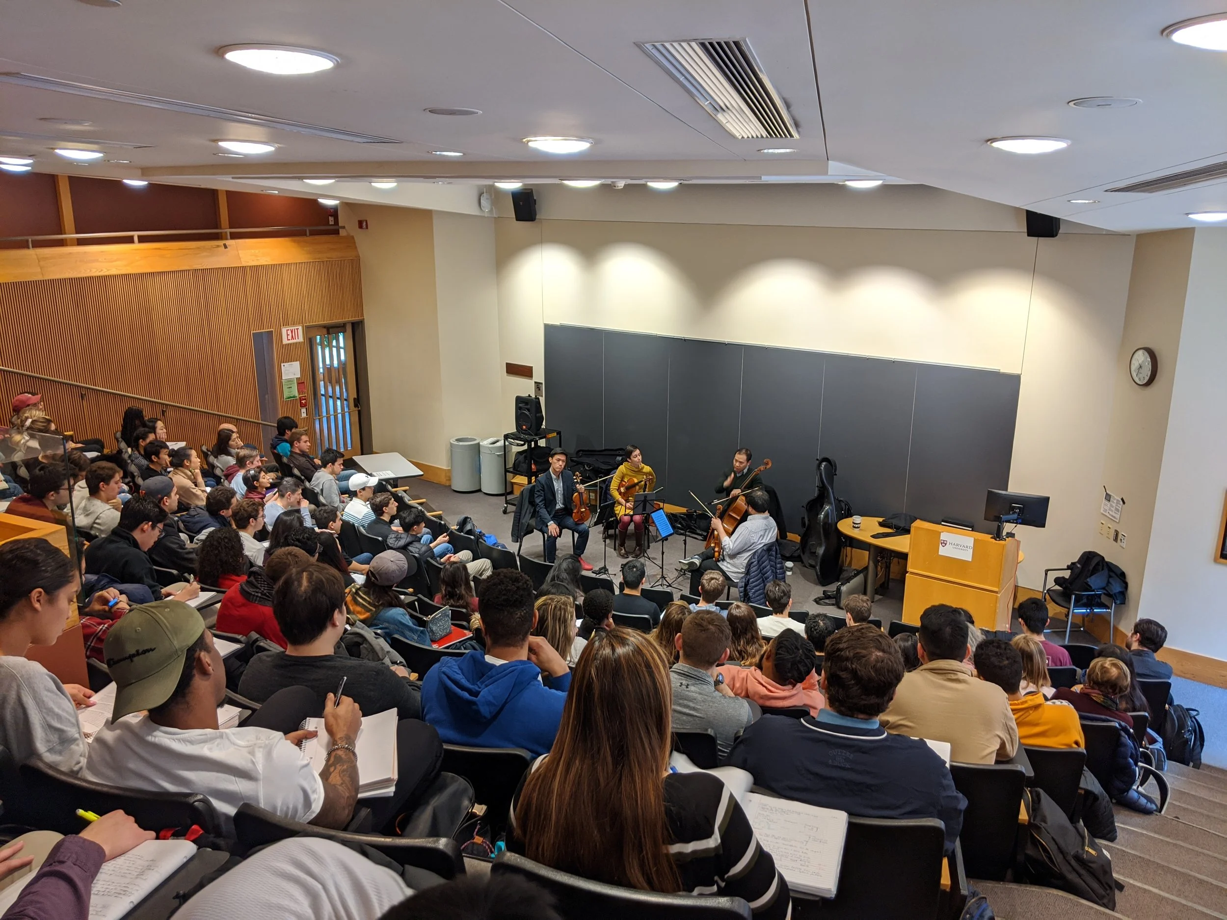 A lecture hall filled with students seated and facing a stage, where musicians are performing with string instruments.