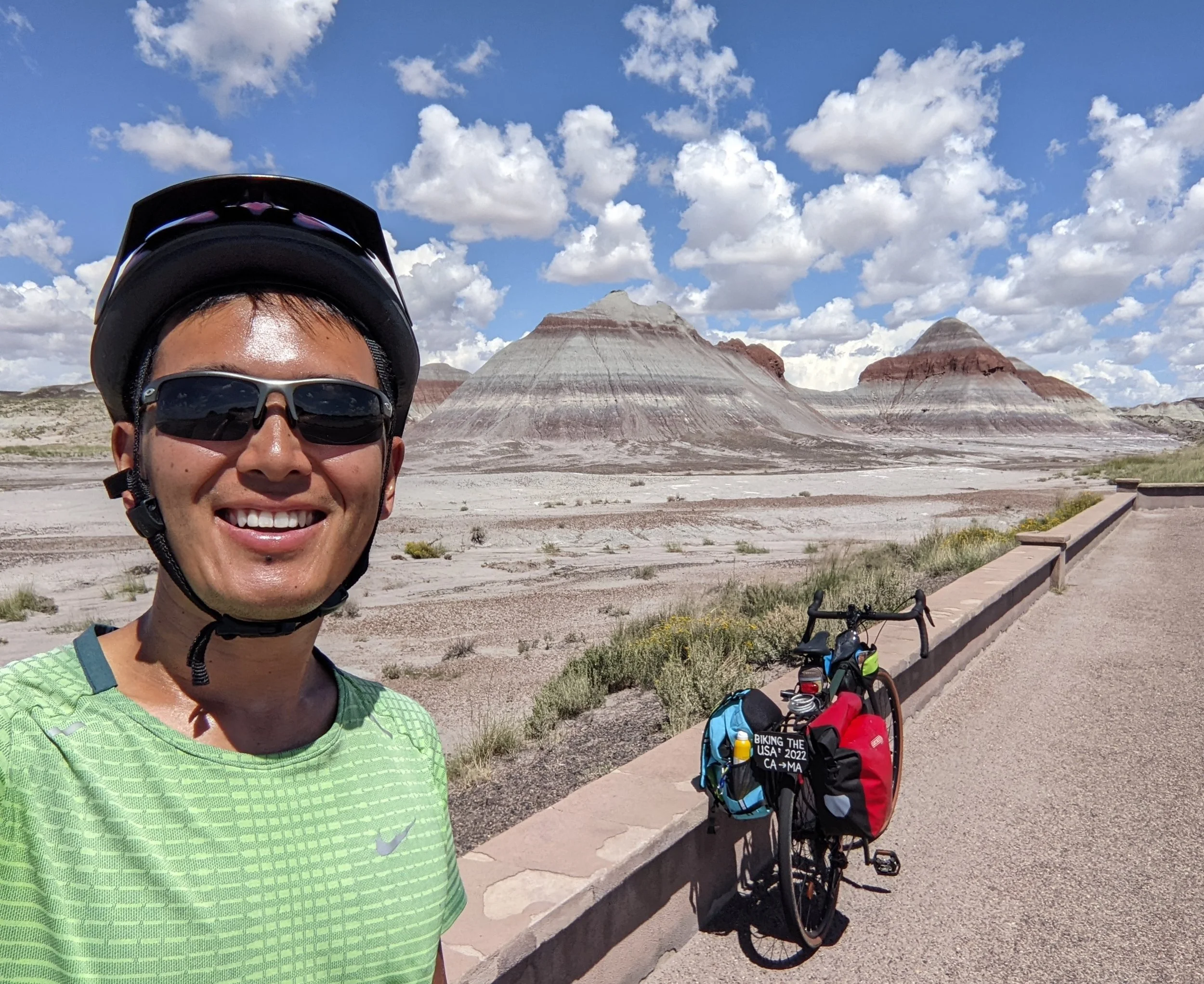A person wearing a green shirt, sunglasses, and a cycling helmet, smiling in front of a desert landscape with colorful buttes and a partly cloudy sky. There is a bicycle with bags and water bottles in the background.