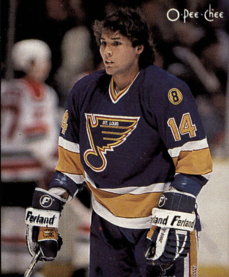 A young man in a blue New York Rangers hockey uniform with the number 14, standing on the ice during a game, with other players and spectators blurred in the background.