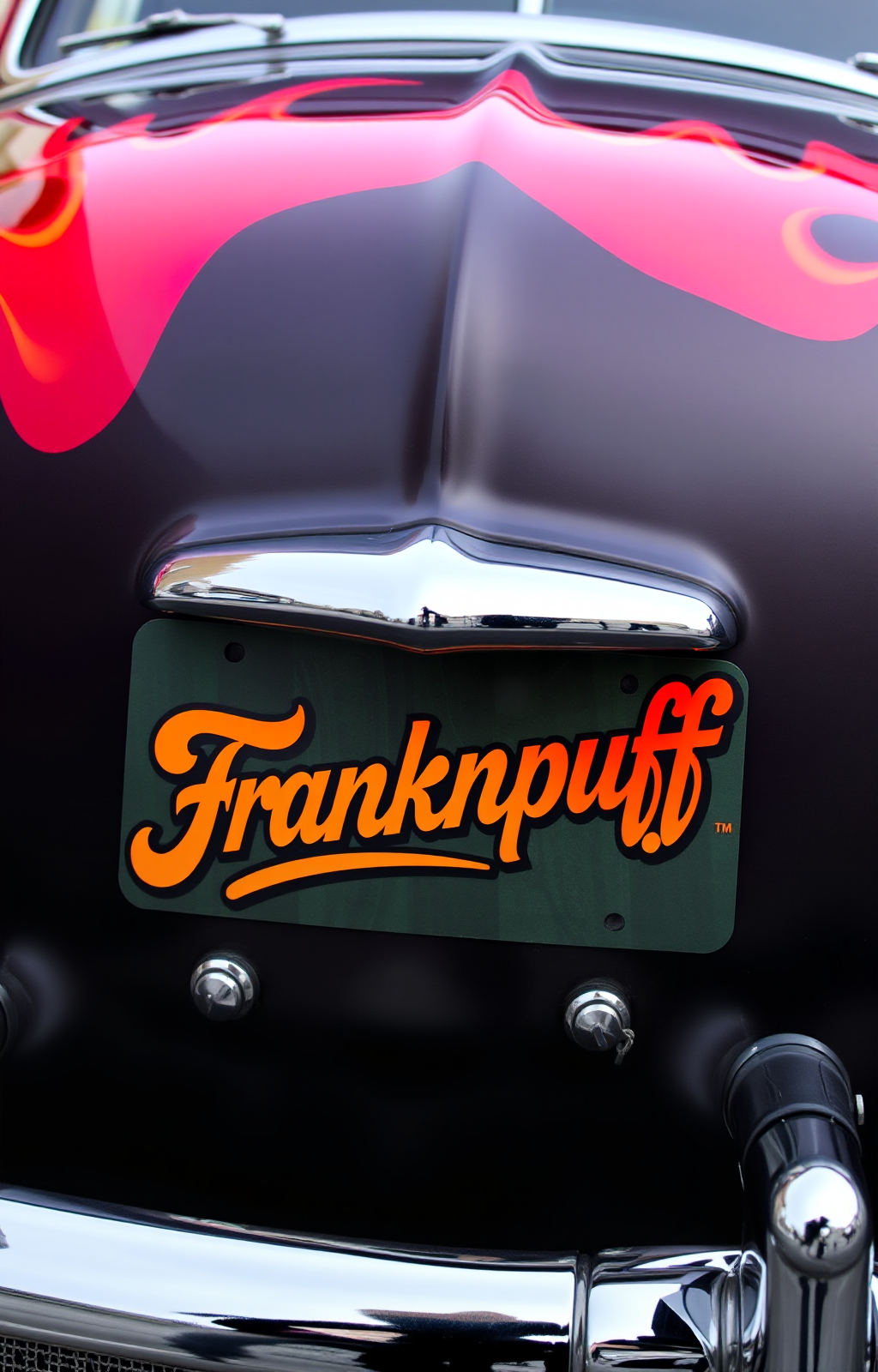 Close-up of a vintage car's front with a license plate that reads 'Franknpuff'. The car has a shiny black hood with pink and yellow flame decals and a chrome bumper.