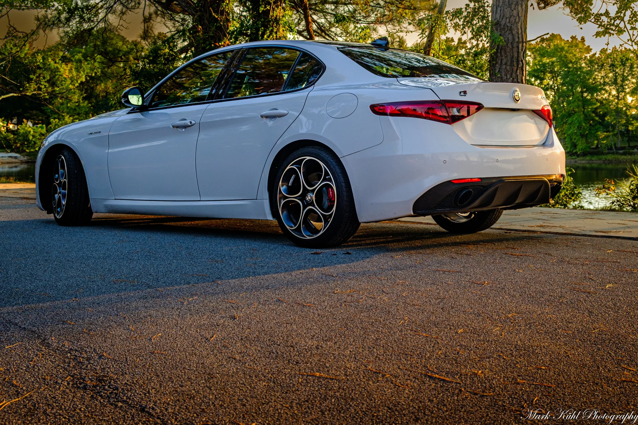 White sedan car parked on a paved surface near trees with green leaves in the background during sunset.