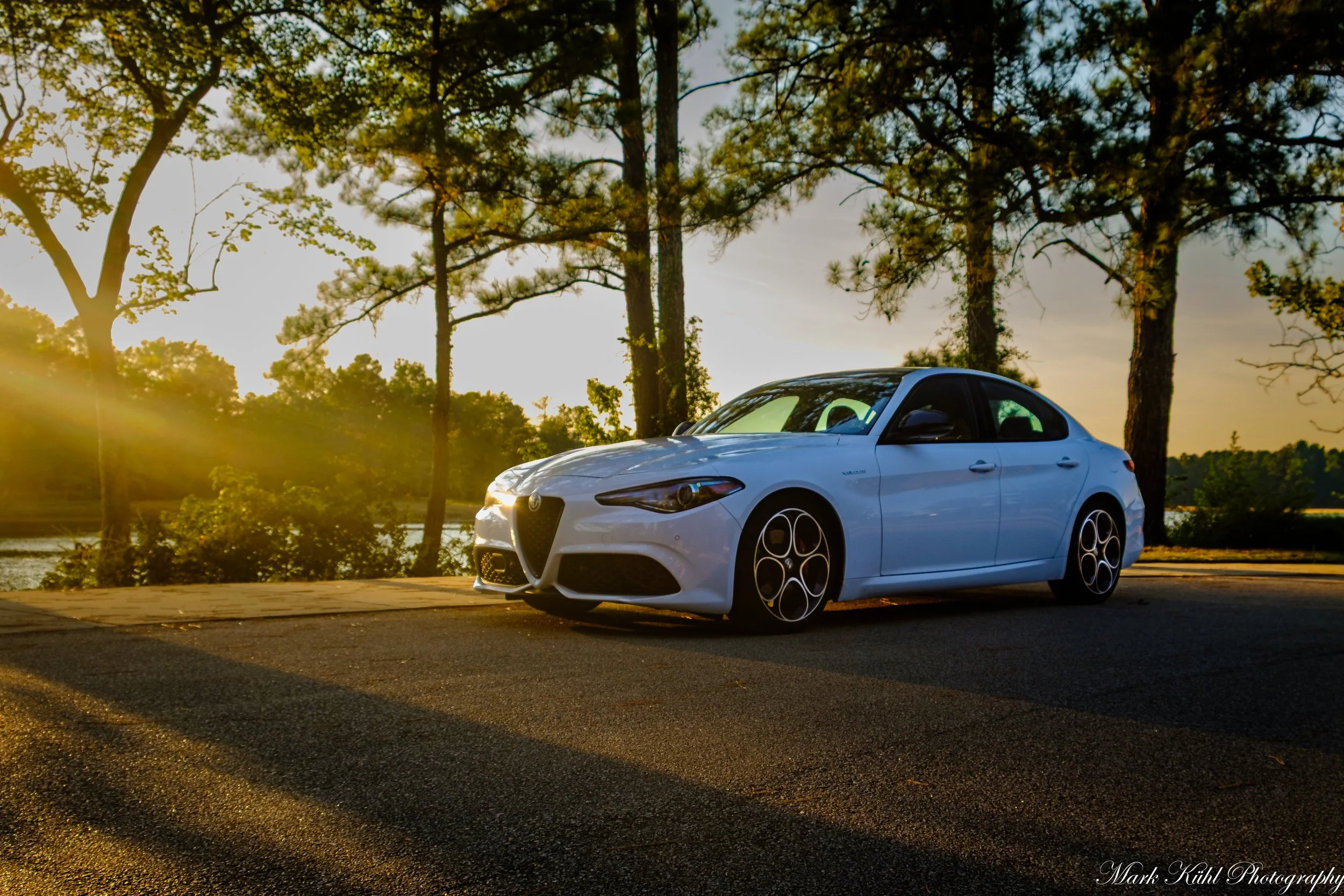 A white sports car parked on a paved road near a lake, with tall trees and a sunset sky in the background.