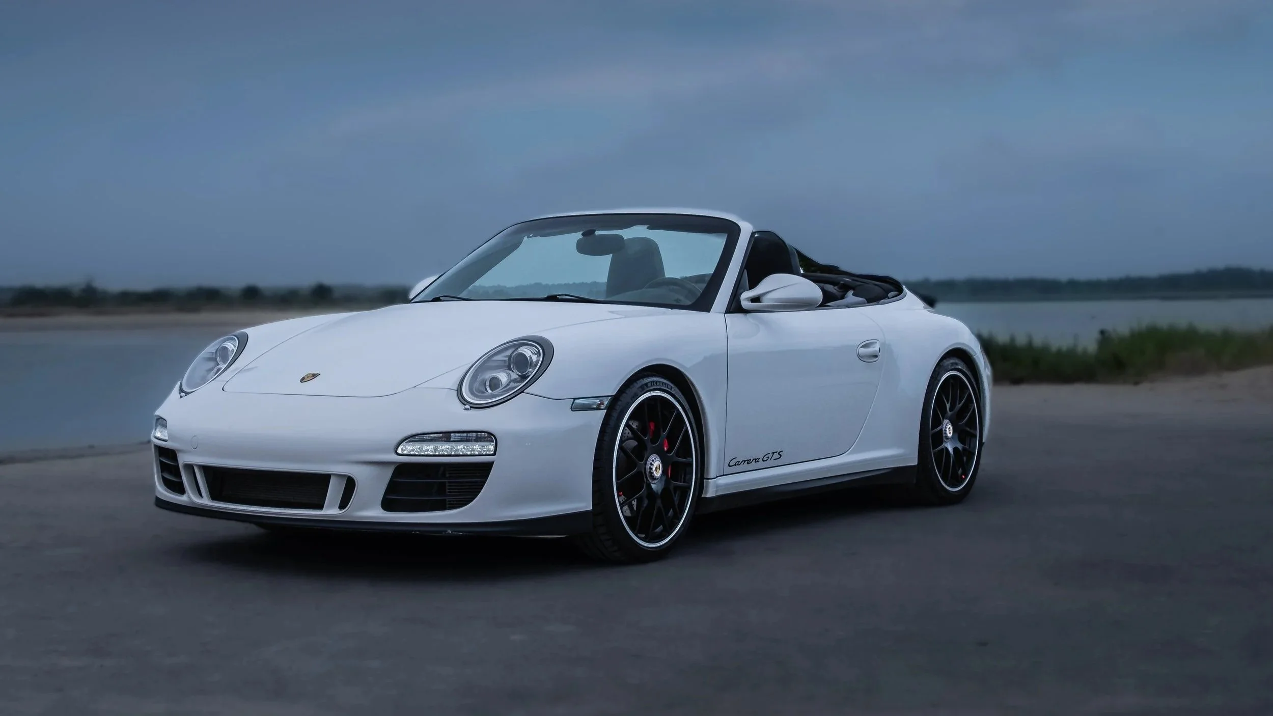 A white Porsche Carrera GTS convertible sports car parked near a body of water under a cloudy sky.