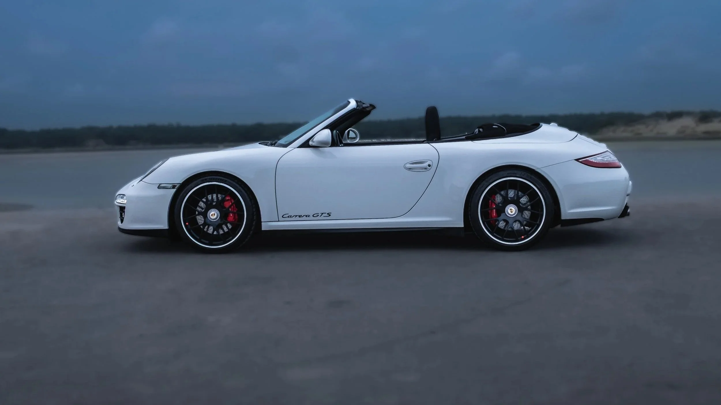 A white Porsche Carrera GTS convertible sports car driving on a coastal road during cloudy weather.