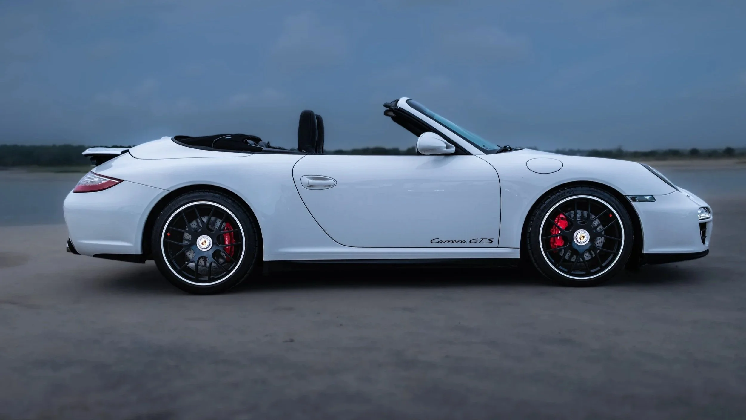 A white Porsche Carrera GTS convertible with its top down, parked on an empty road under a cloudy sky.