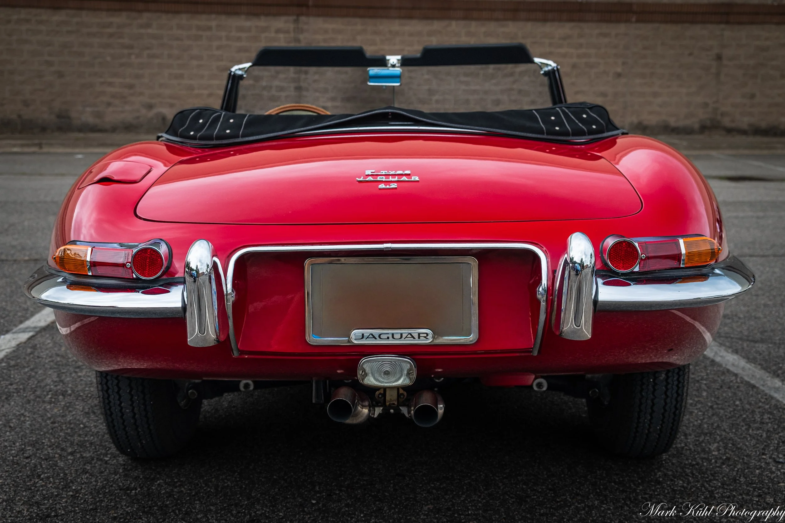 Rear view of a classic red Jaguar convertible sports car parked on pavement, featuring round taillights, chrome details, dual exhaust pipes, and a black soft top folded back.