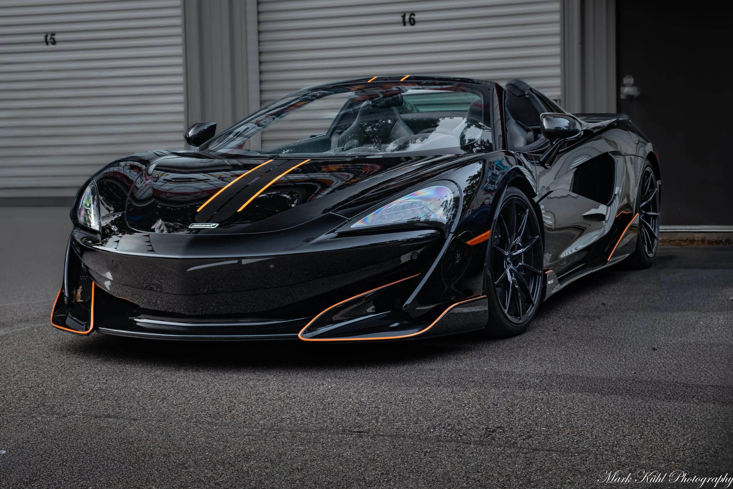 A sleek, black sports car with orange accents parked in front of gray metal storage units, reflecting its surroundings.