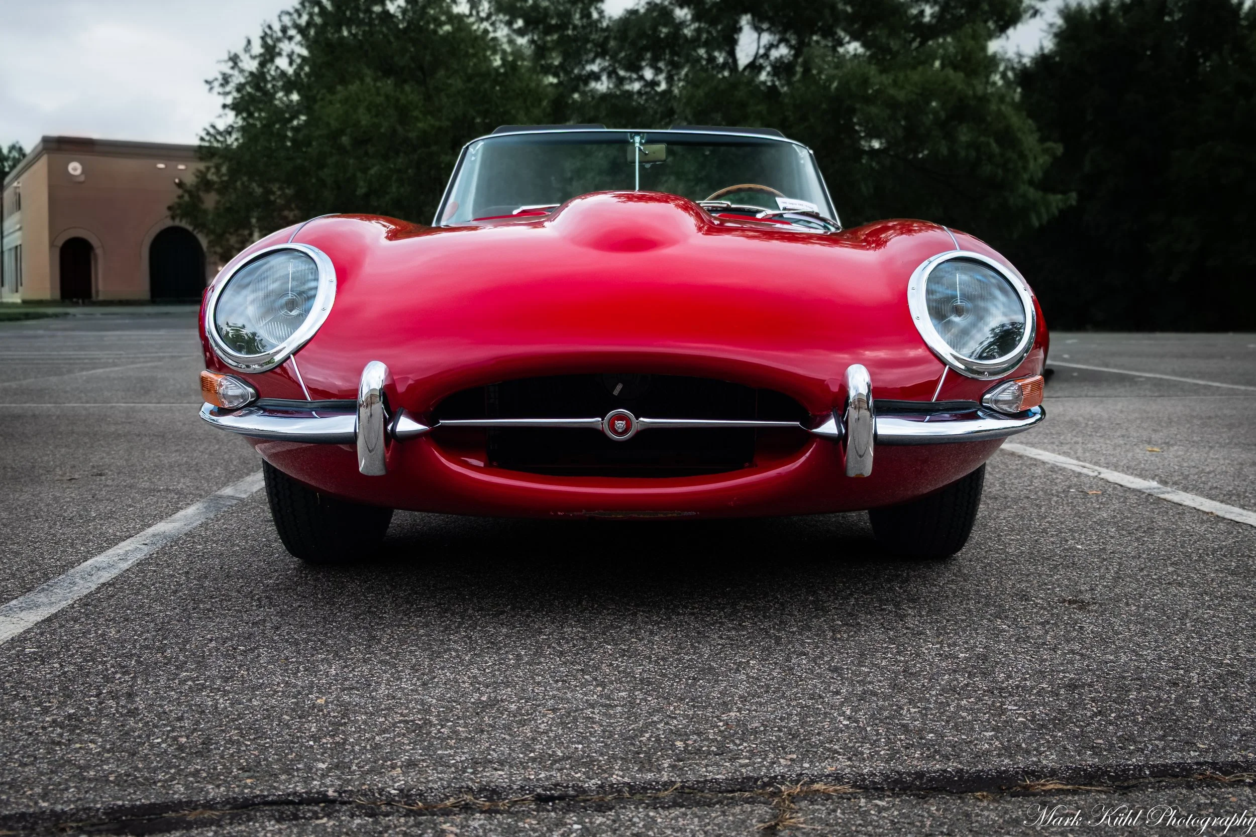 Front view of a shiny red vintage sports car parked in an empty parking lot with a brick building and trees in the background.