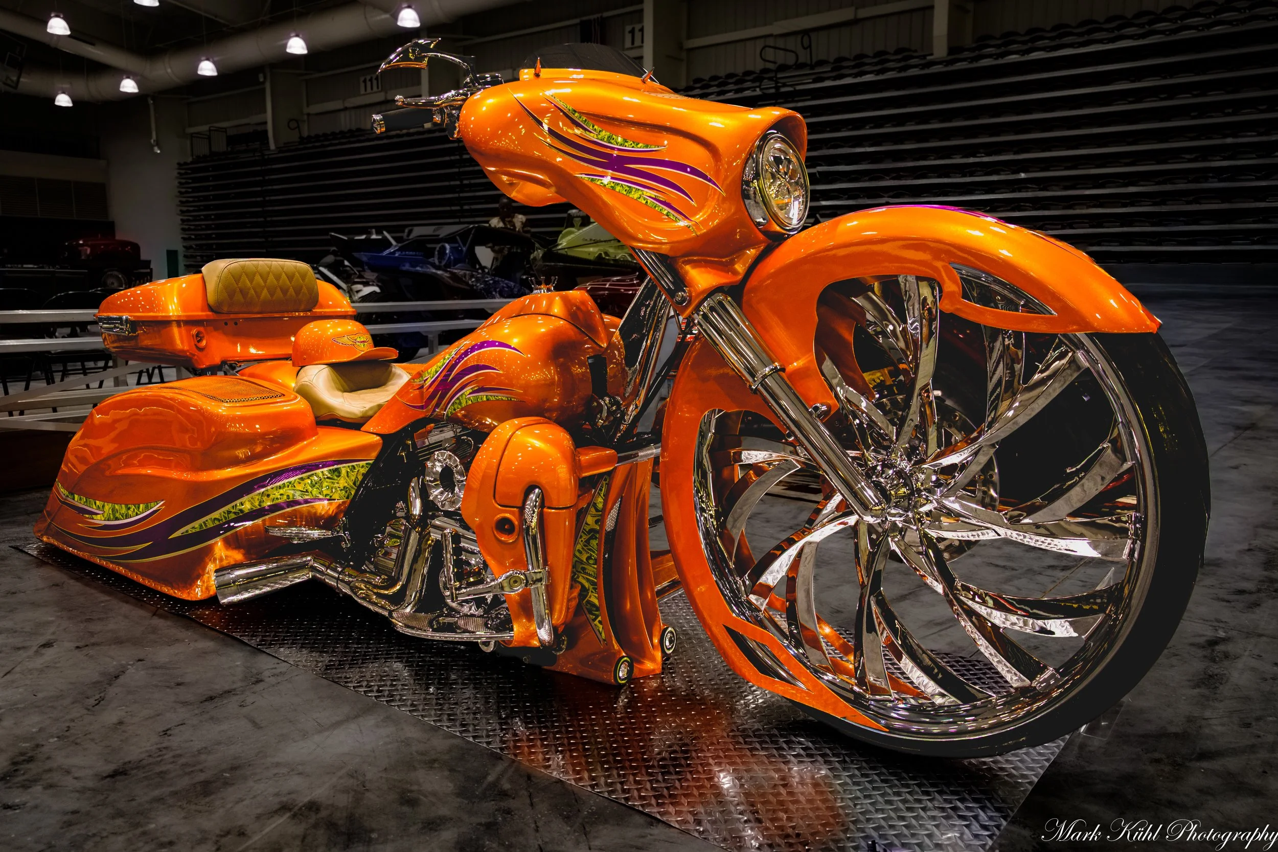 A custom bright orange motorcycle with purple and yellow flame decals, chrome wheels, and a matching orange helmet with decals, displayed indoors at an exhibition.