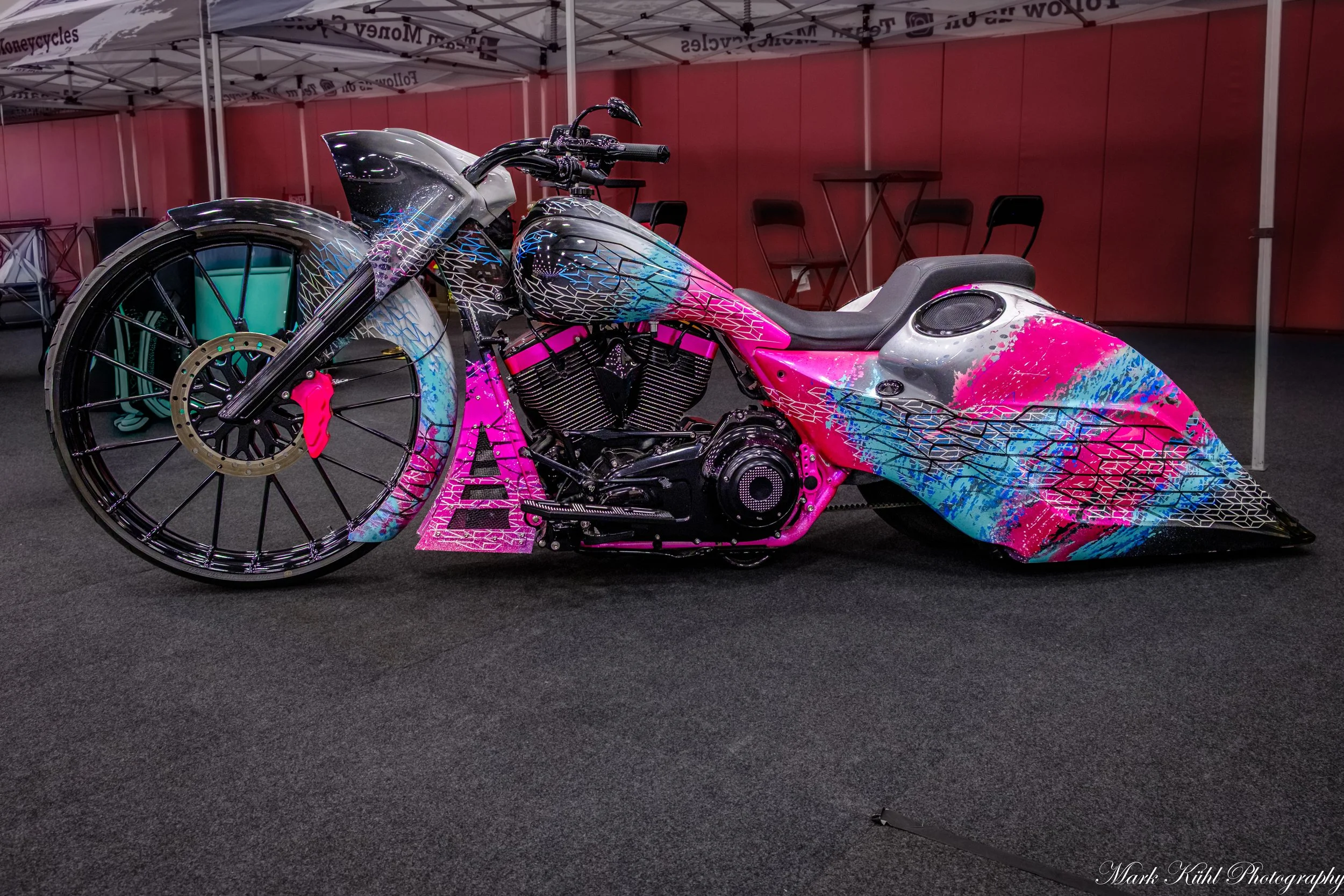 A custom motorcycle with a pink, blue, and black paint job, on display at an indoor exhibit, behind a red wall and under a white canopy.