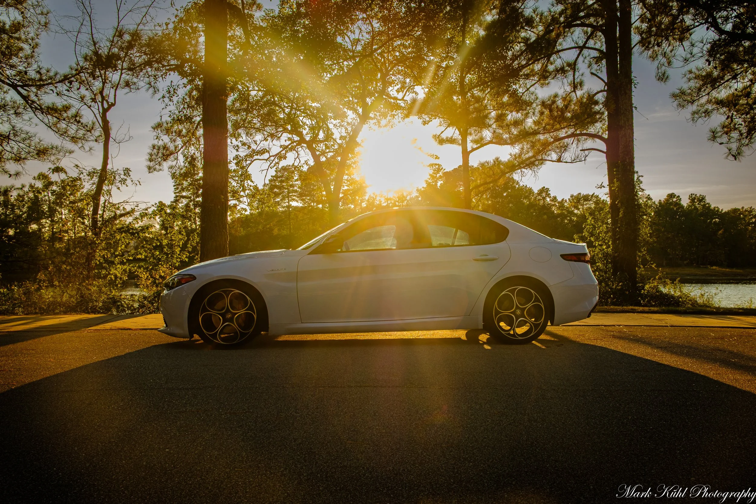 A white coupe car parked on a road near a body of water with tall trees and green foliage in the background. The sun is setting or rising, creating a bright glare and sun rays through the trees.