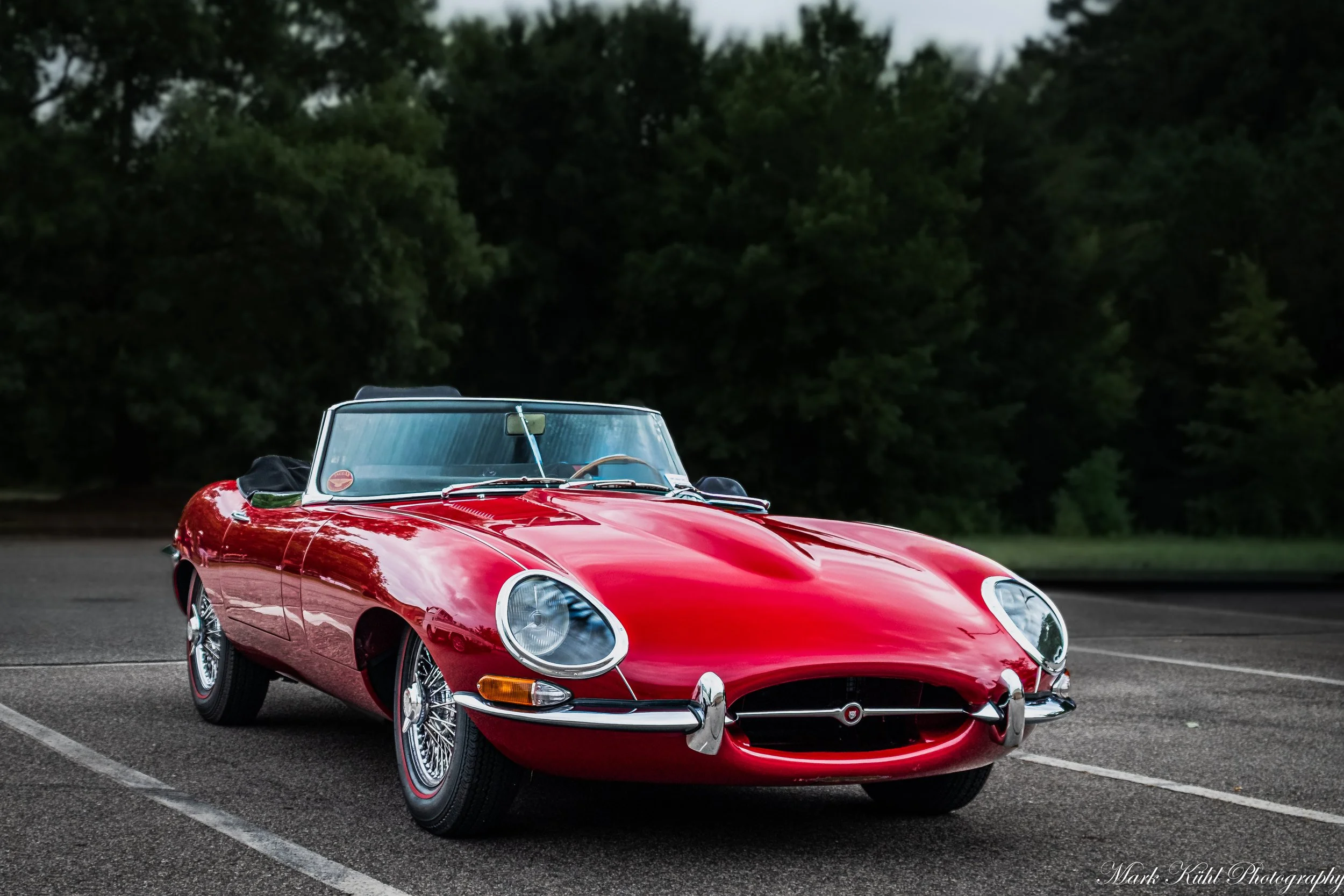 A vintage red sports car parked in an outdoor lot surrounded by trees.