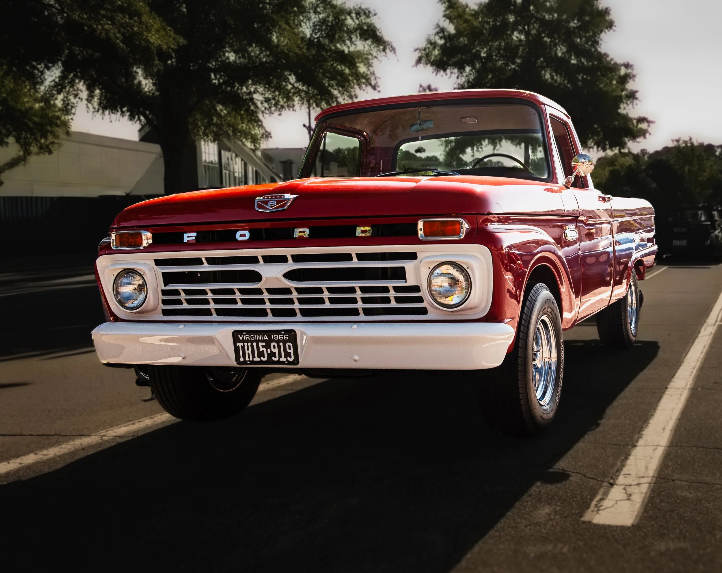 A red vintage Ford pickup truck parked on the street with trees in the background and a Virginia license plate that reads 'TH15-919'. The truck has a chrome side mirror and an emblem on the front hood.