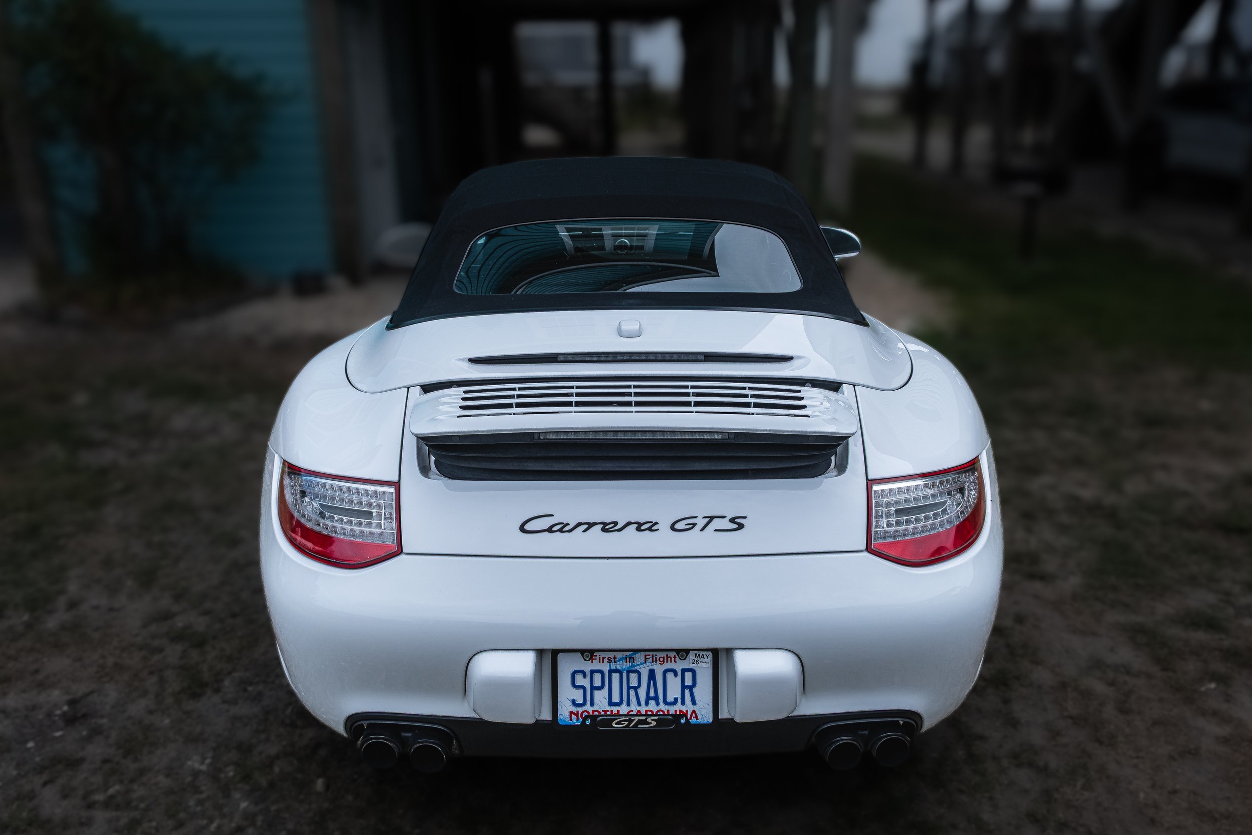 White Porsche Carrera GTS convertible car parked outside with North Carolina license plate reading 'SPORAC.' The car is on a dirt or gravel surface, with a wooden fence and some bushes in the background.