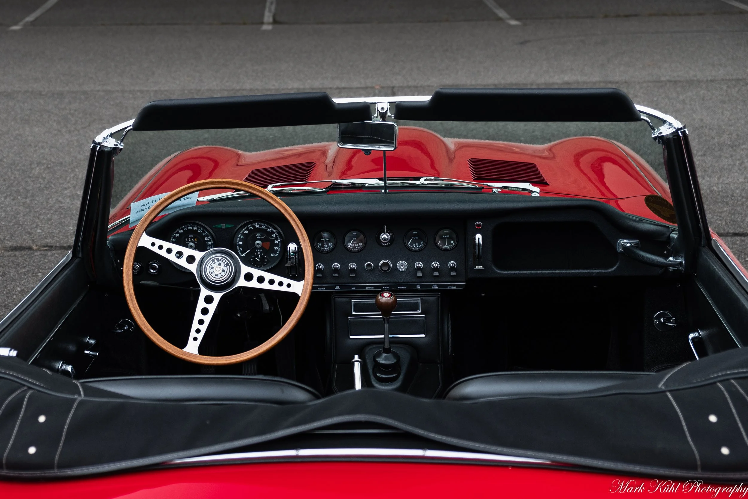 The interior of a red vintage sports car with a tan wooden steering wheel, analog dashboard gauges, and a manual gear shift, parked on a paved surface.