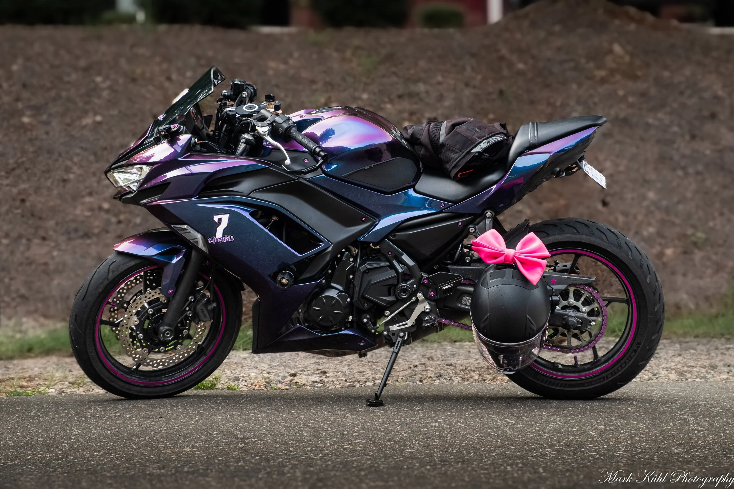 A purple and black sport motorcycle with pink accents and a pink bow on the rear, parked on a street with a dirt mound in the background.
