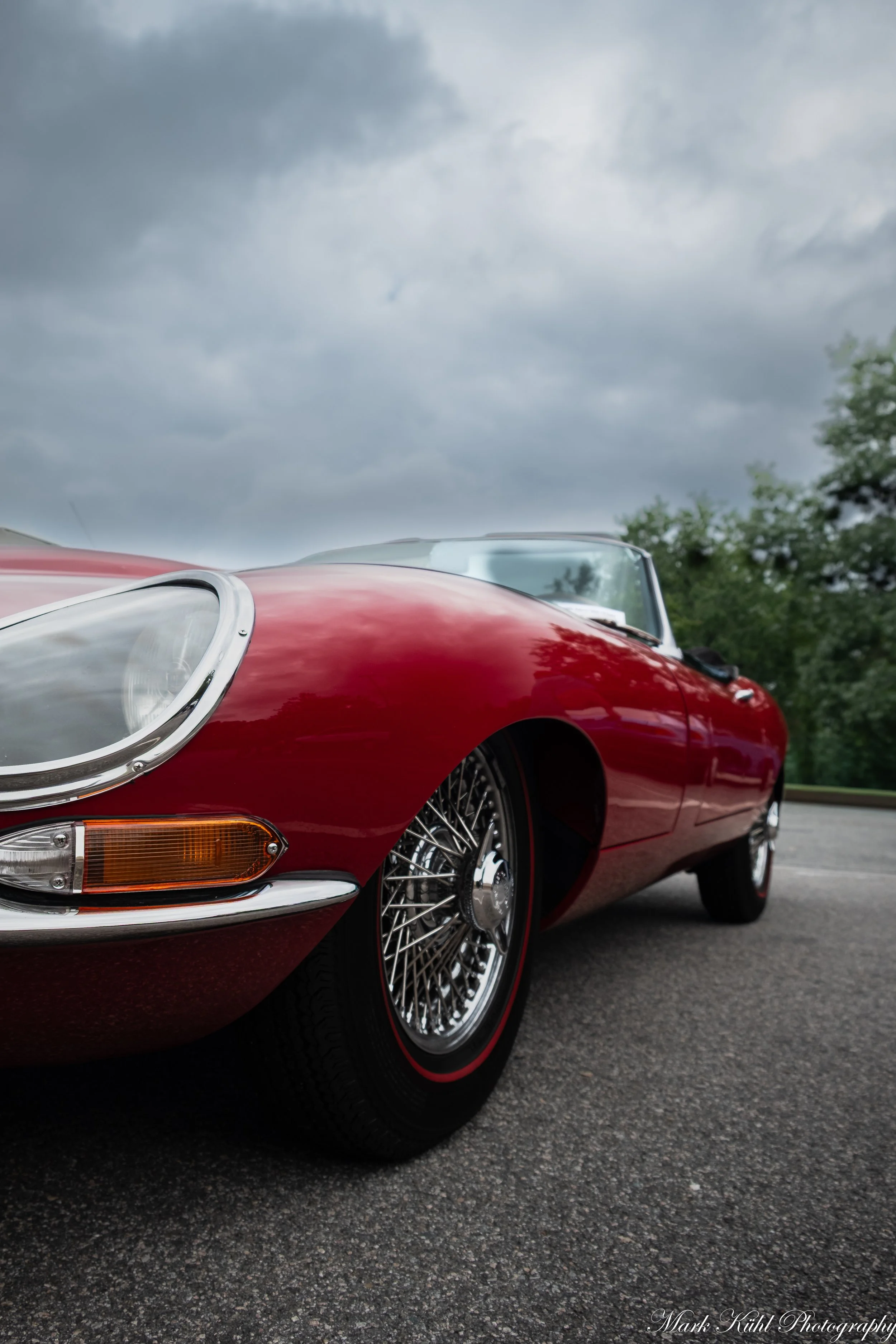 Close-up of a vintage red sports car with wire-spoked wheels parked outdoors on an overcast day, with trees visible in the background.
