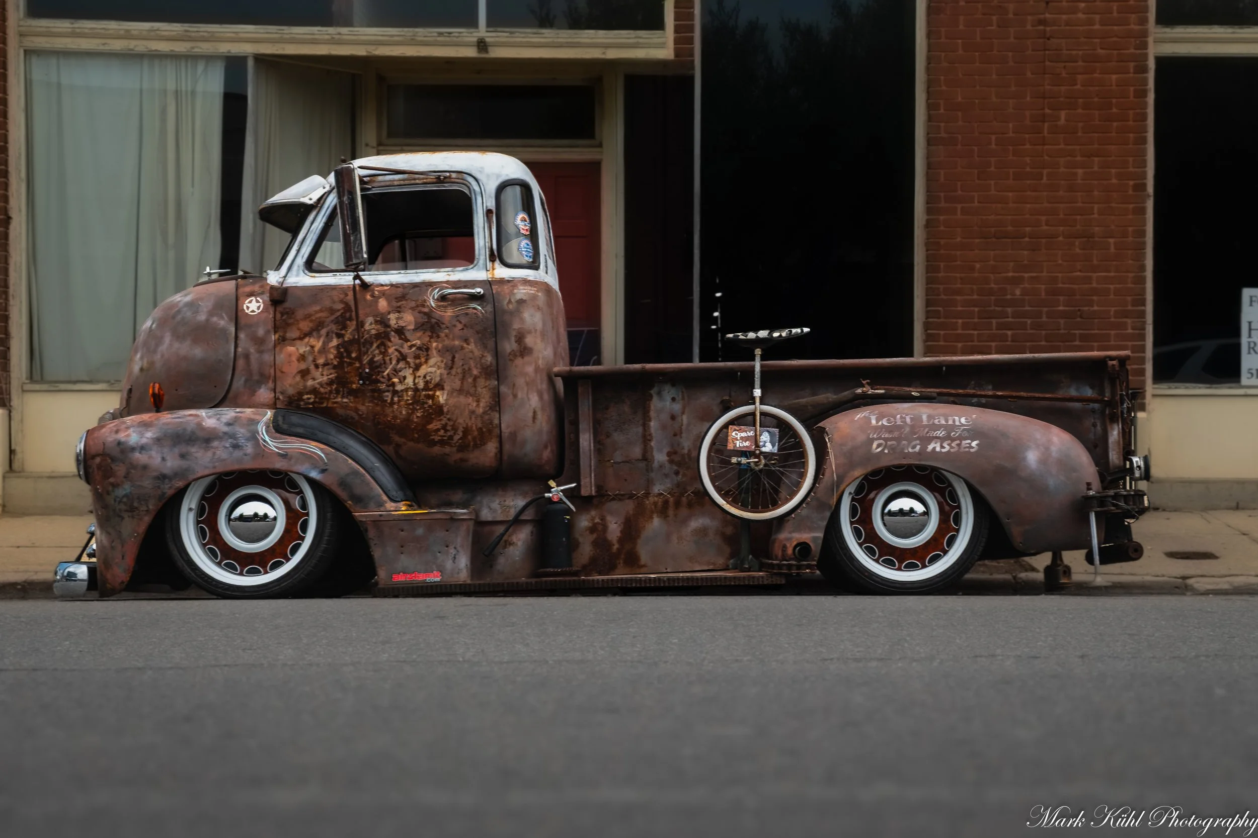 An old rusty pickup truck parked on the street in front of a building with large windows and brick walls. The truck has a bicycle mounted on the side and various stickers on the window.