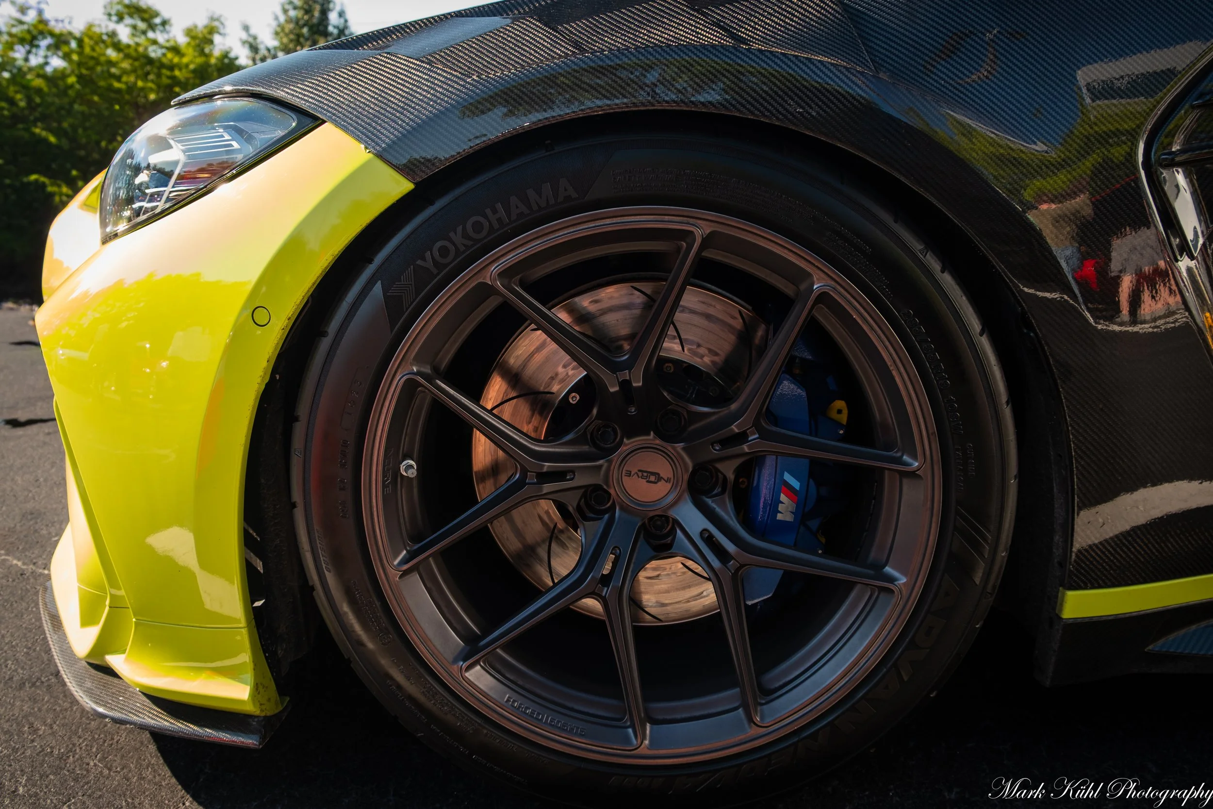 Close-up of a sports car's front left wheel with a bronze-colored rim and a Yokohama tire, partially covered by a carbon fiber fender, with the car's headlight and yellow body visible.
