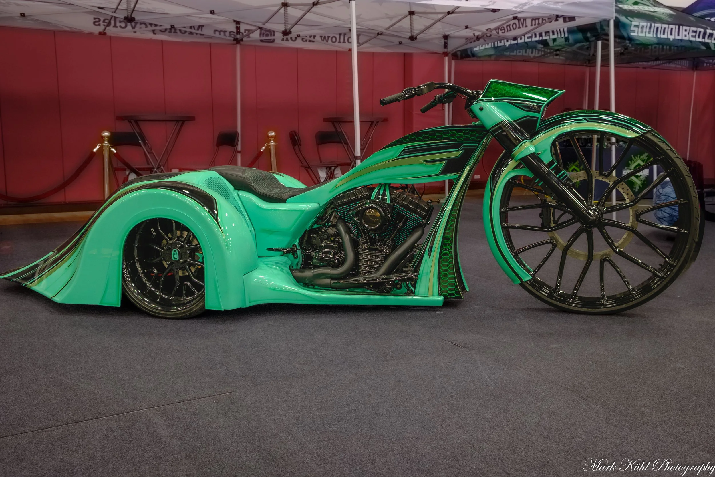 A futuristic green motorcycle with sleek, aerodynamic design and large black wheels is displayed indoors, under white tents, with red walls and chairs in the background.