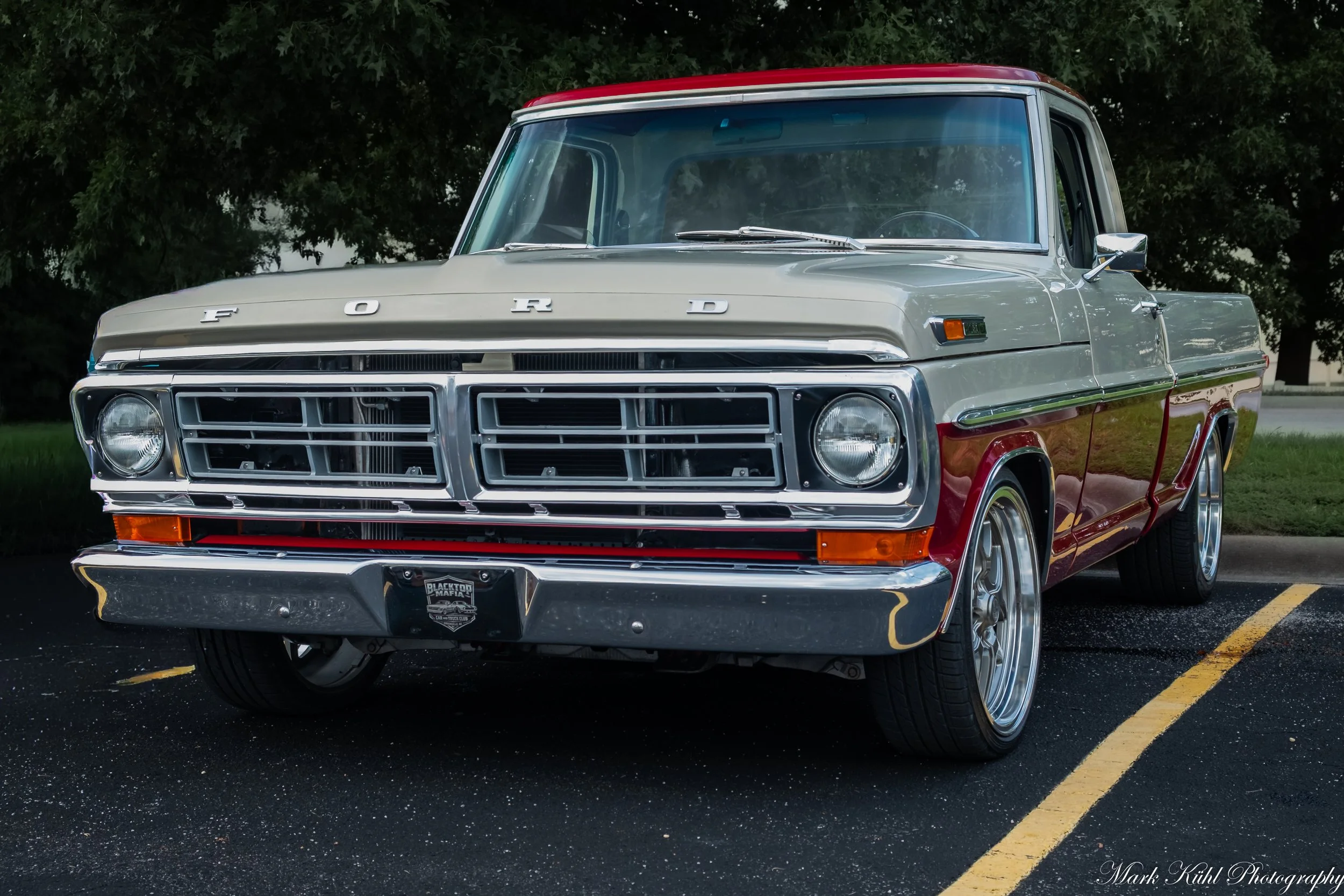 A vintage Ford pickup truck parked in a parking lot with a background of trees, featuring a gray and red color scheme and chrome accents.