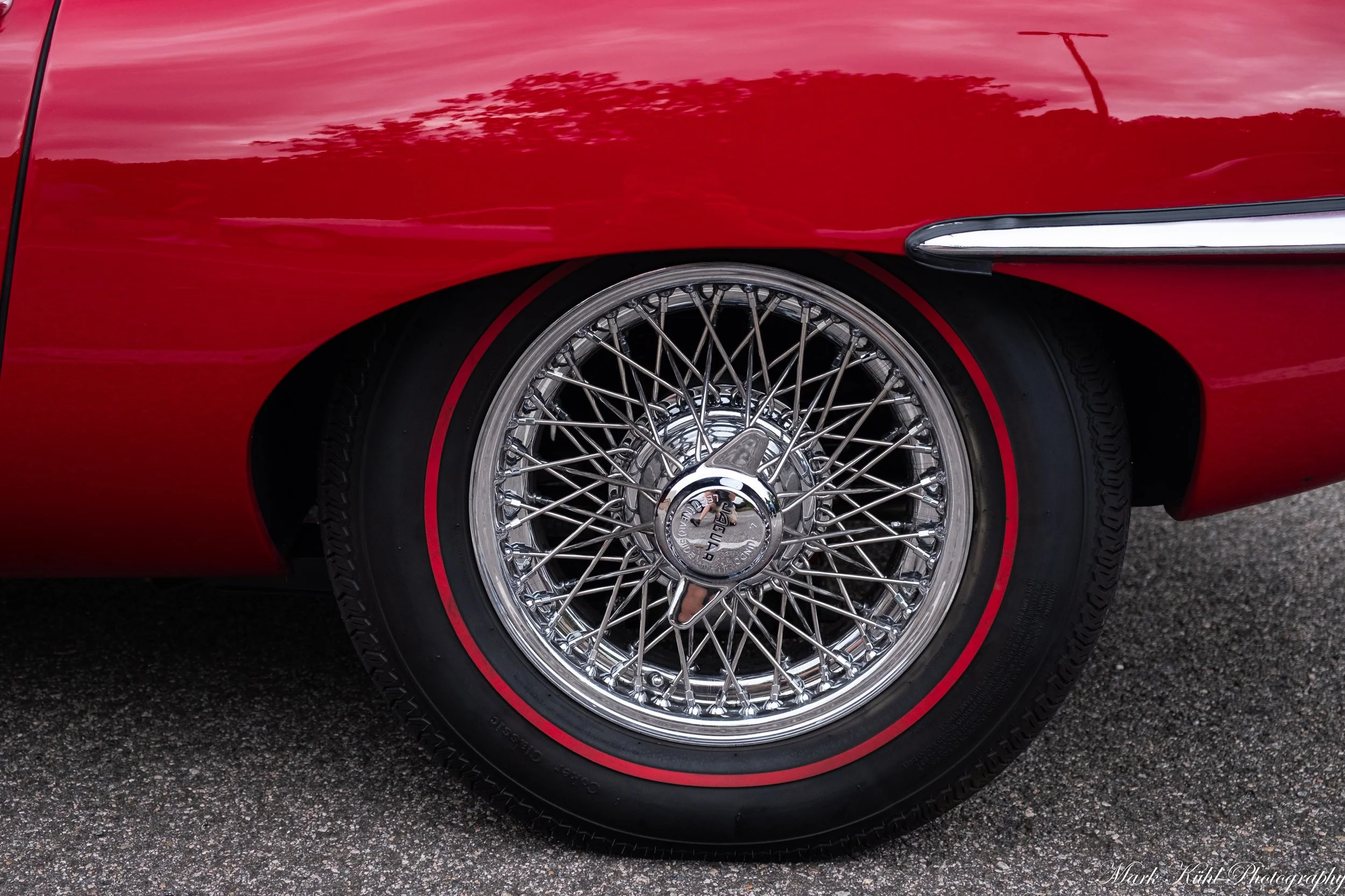 Close-up of a classic red sports car, focusing on the front right wheel with intricate wire spokes and a silver hubcap, set on a paved surface.