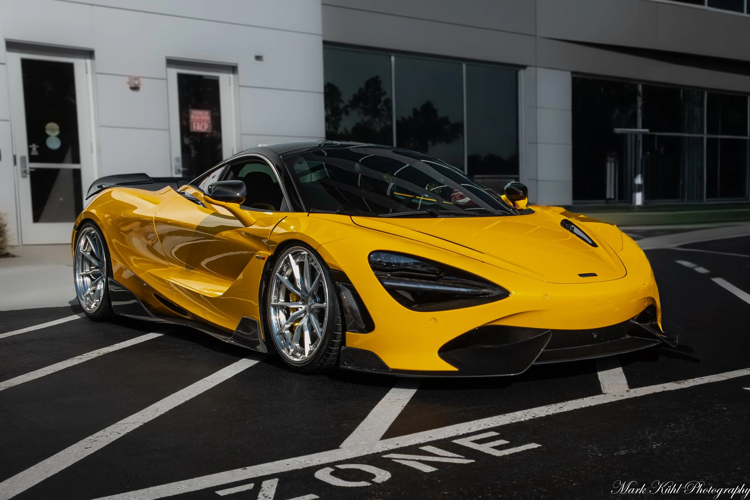 A yellow sports car parked in a parking lot in front of a modern building with large glass windows and white doors.