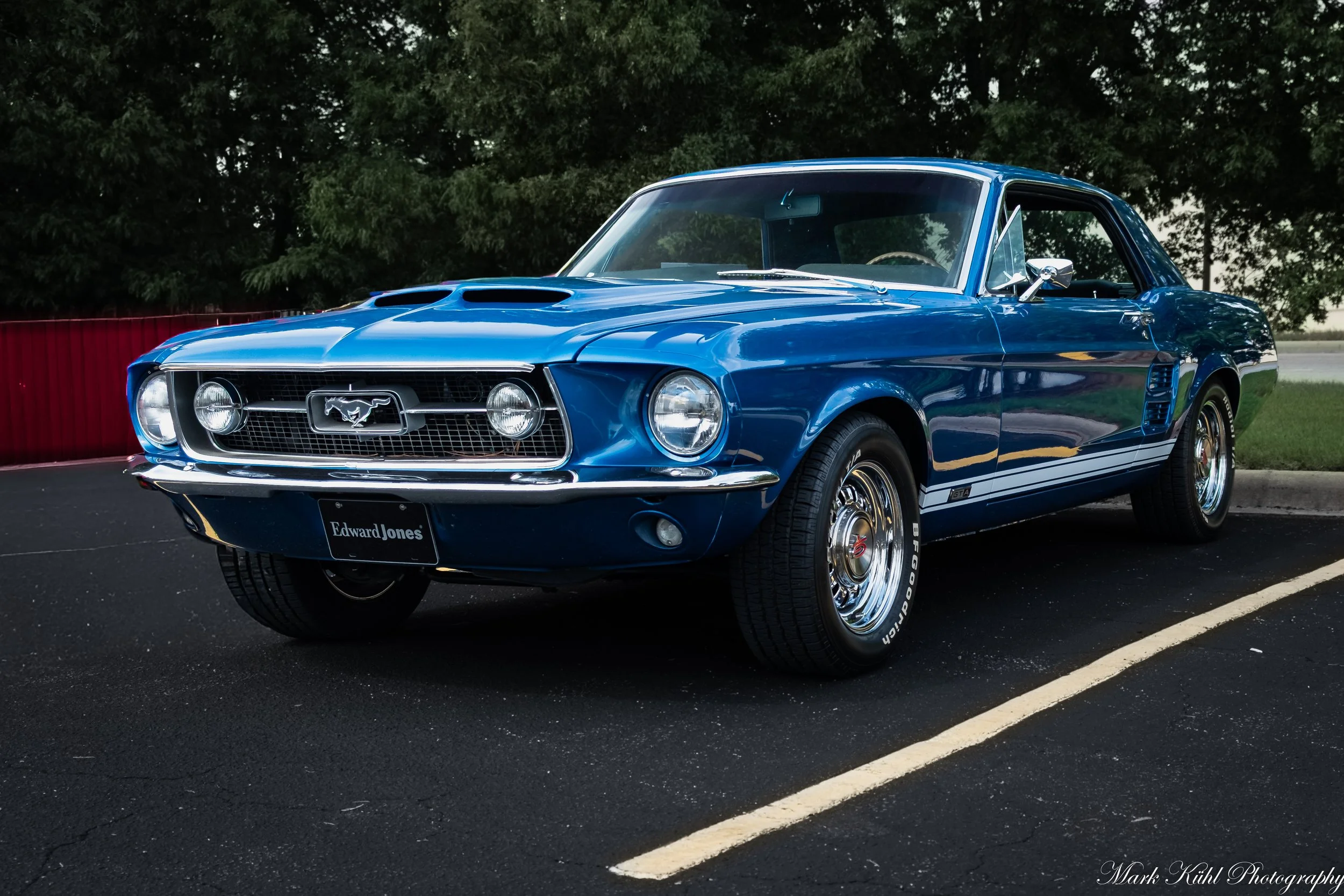 A bright blue vintage Ford Mustang parked on a paved parking lot with trees in the background.