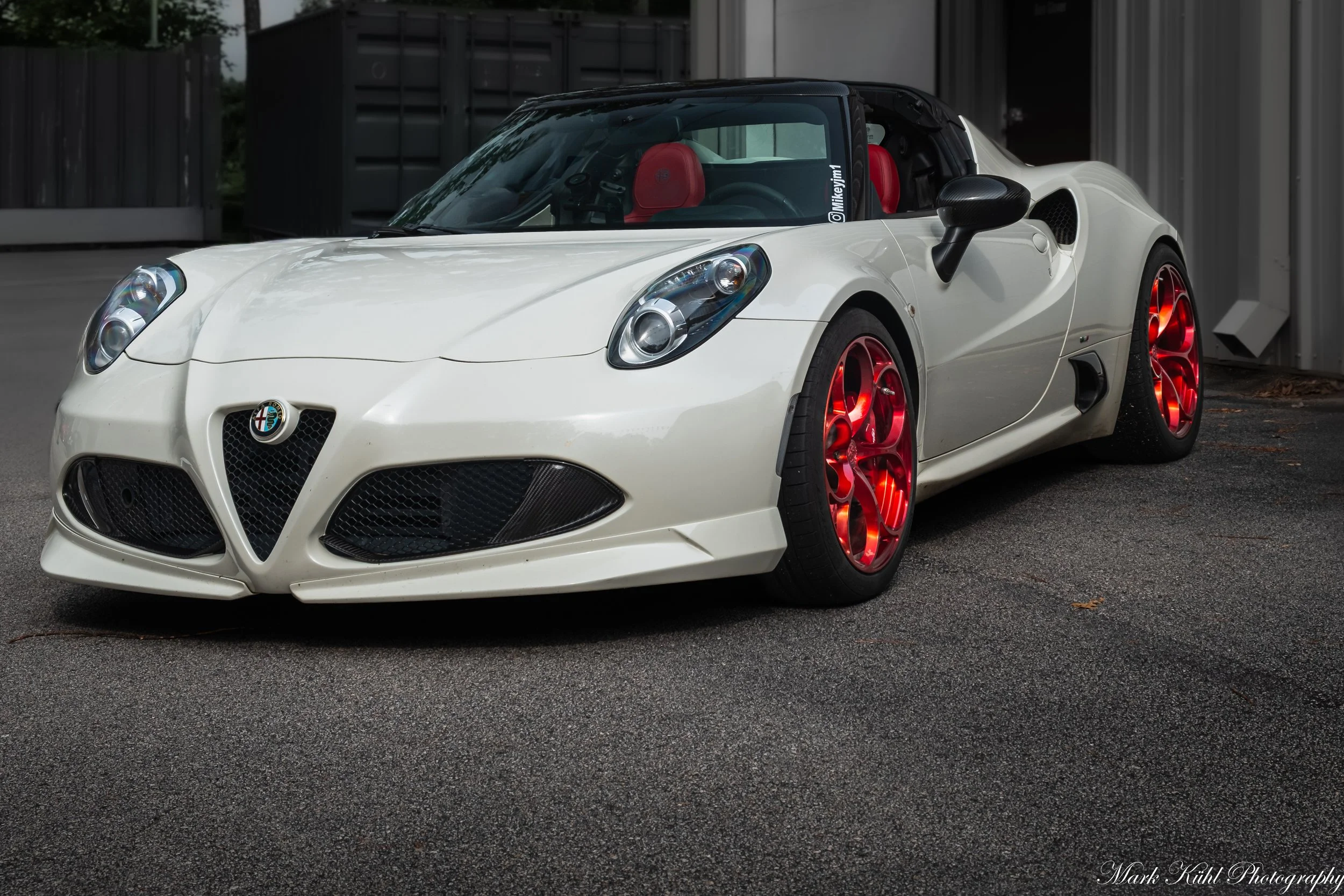 A white Alfa Romeo sports car with red rims parked outdoors in front of a gray fence, with a black convertible roof, red interior seats, and a sticker on the windshield with the text '@Mikevykim'.