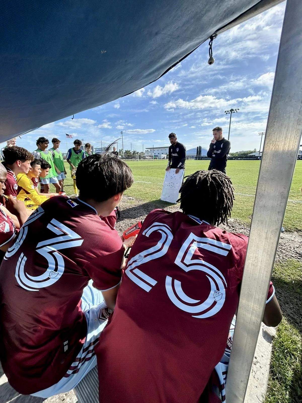 Soccer team listening to coaches during a sideline strategy session on a sunny day at a sports field.