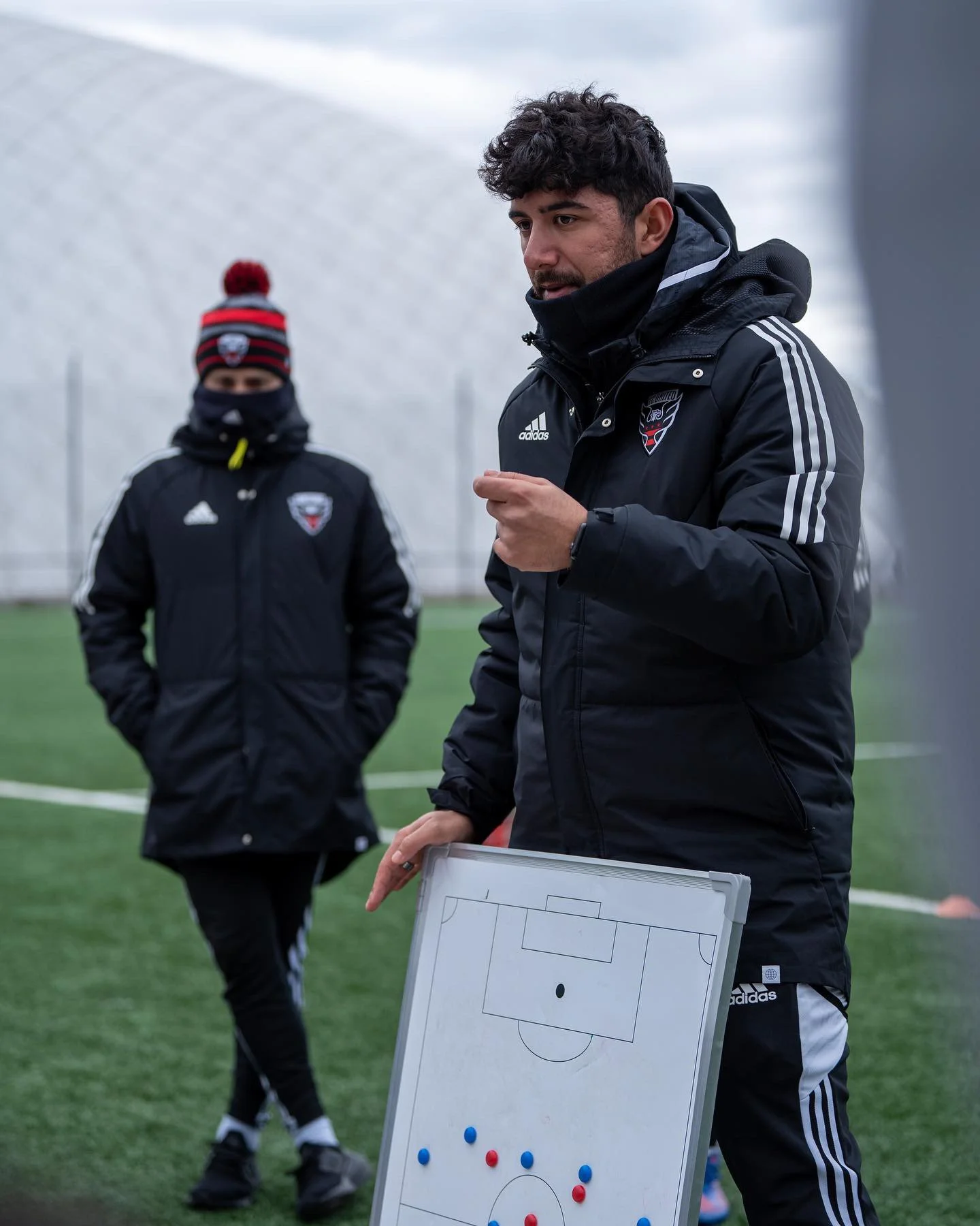 Two soccer coaches on a field, one holding a tactical whiteboard, wearing black jackets with Adidas logo, during a training session in cold weather. One coach is explaining a strategy, the other is listening with his hands in his pockets. Both are dressed for cold weather, with the background showing a large, white, dome-shaped structure.
