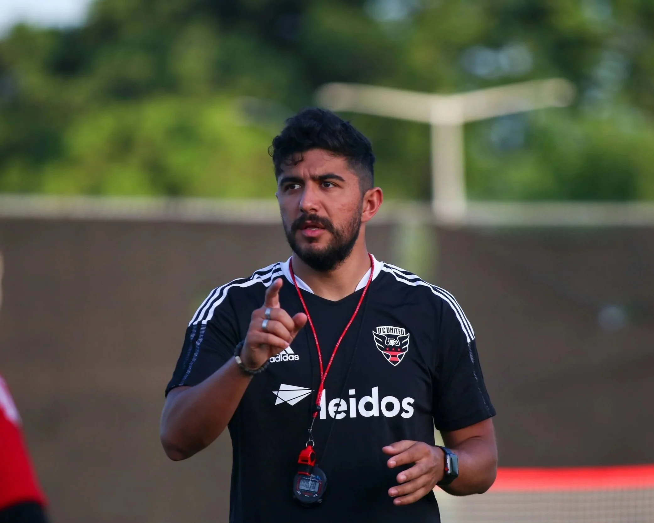 A man with dark hair and a beard, wearing a black sports shirt with white stripes and the logos 'adidas' and 'leidos,' is running outdoors during daytime. He has a red whistle around his neck and a GPS watch on his left wrist, and he appears to be talking or giving instructions as he runs. The background shows green trees and a building.