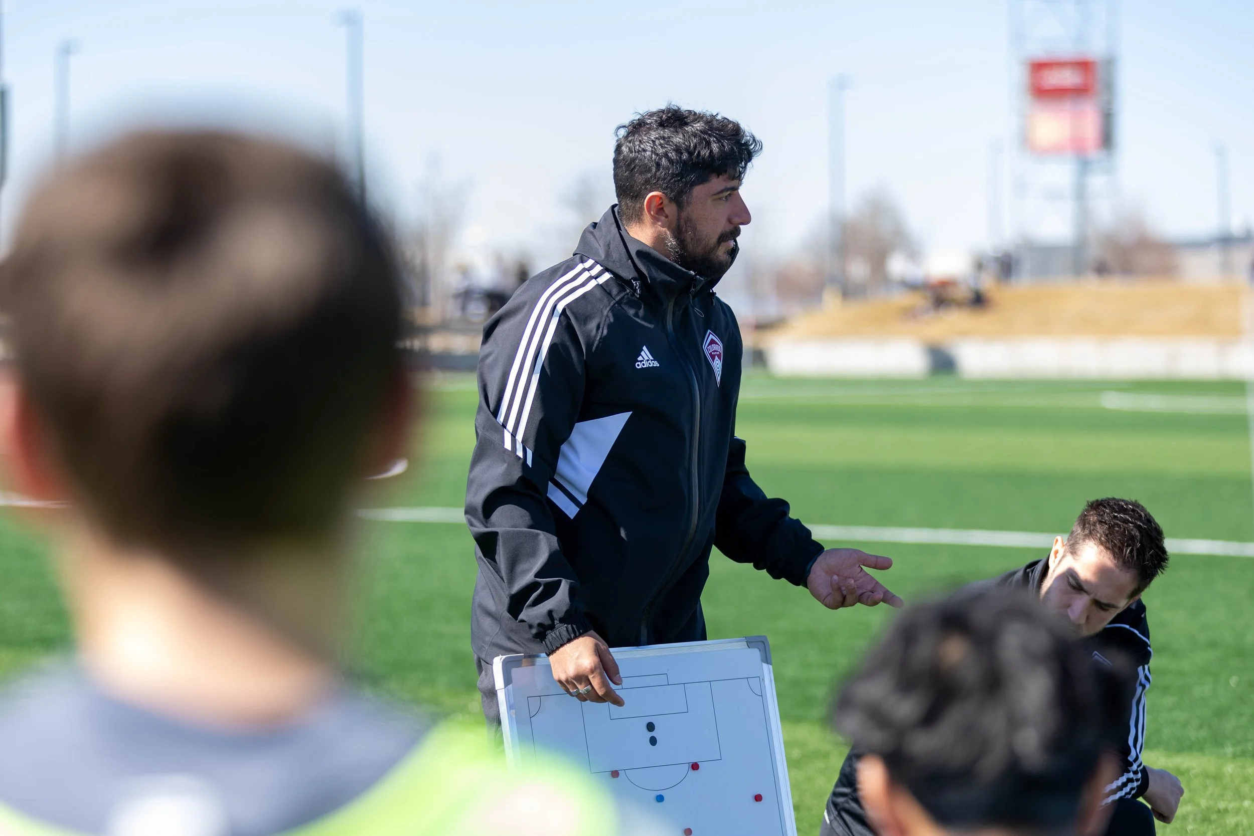 A man with dark curly hair and a beard, wearing a black Adidas sports jacket, is coaching a soccer team on a field during daytime. He is holding a whiteboard with a soccer field diagram and is speaking to the players, who are sitting and listening attentively.