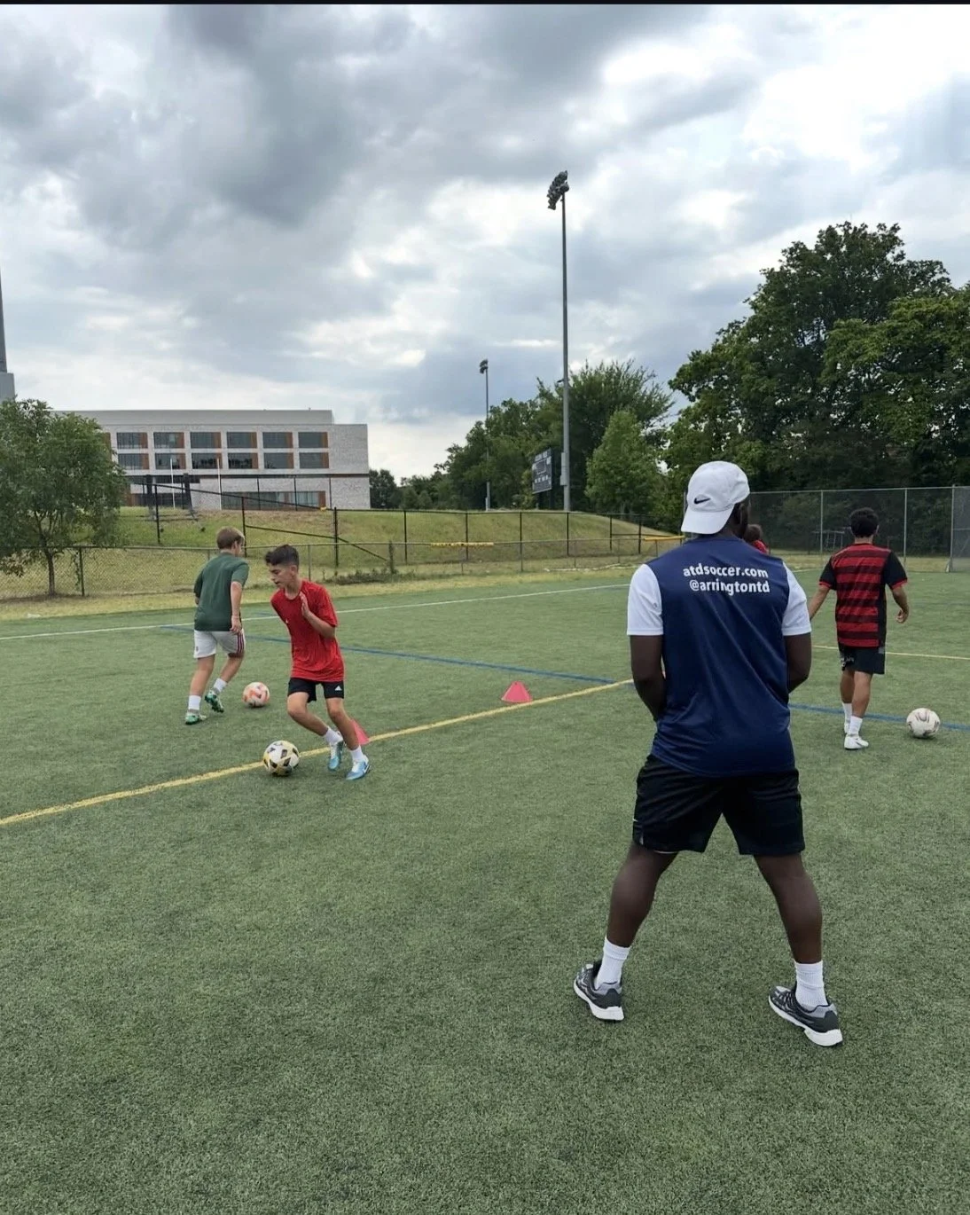 Soccer coach supervising young players during practice on a field with cloudy skies, trees, and a building in the background.