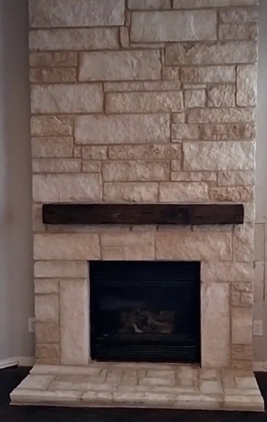 Fireplace made of beige stone with a dark wooden mantel and a black fireplace insert, set against a stone wall with a stone hearth.