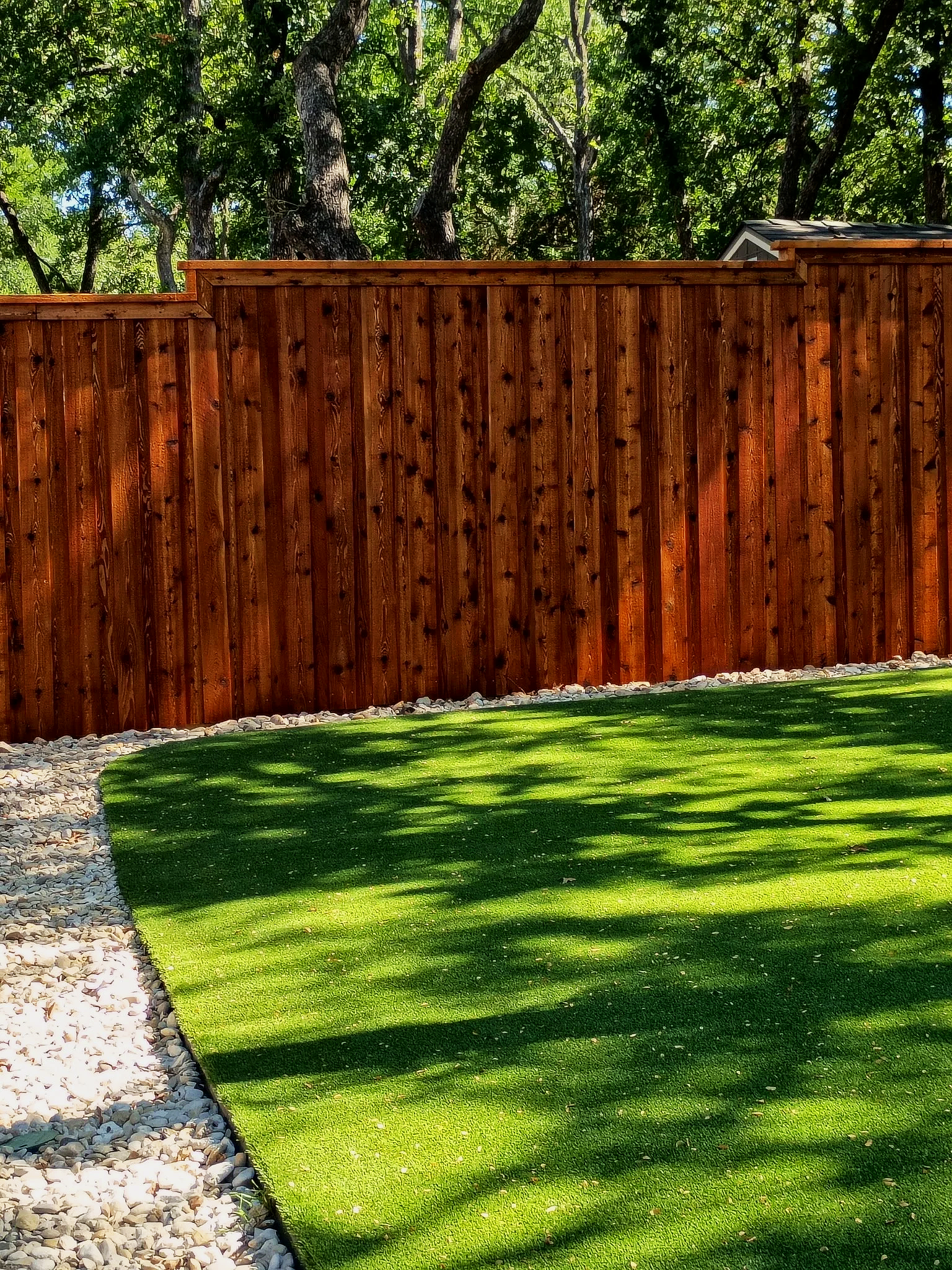 A backyard with a tall wooden fence, a well-maintained green lawn, a gravel border, and trees in the background.