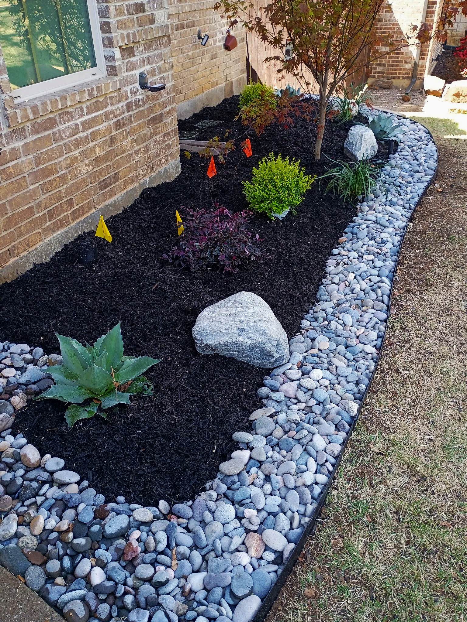 A brick house exterior with a landscaped garden bed along the wall. The garden has dark mulch, small plants, a small tree, and large decorative rocks. There is also a border of smooth, rounded gray and white stones surrounding the garden bed.