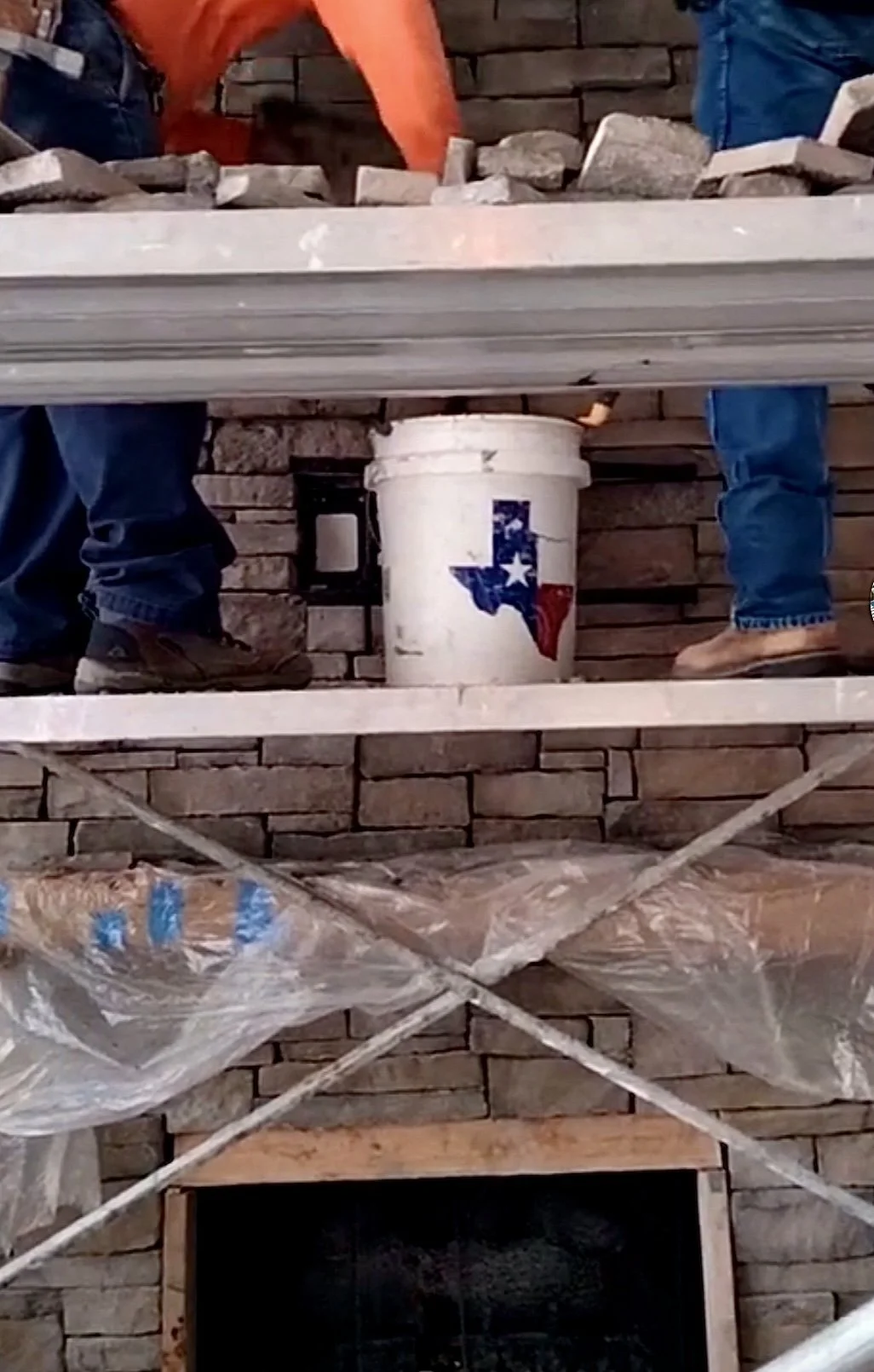 Construction site with brick wall, scaffolding, and workers. A bucket with Texas flag design is placed on a wooden plank.