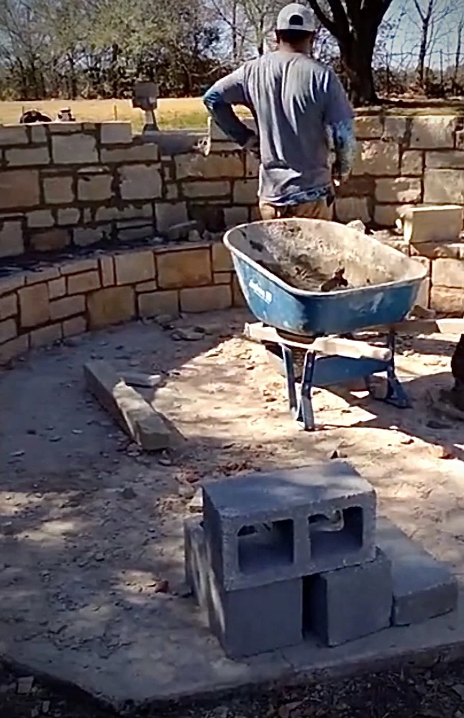 A person working on building a brick wall outdoors, with a blue wheelbarrow and cinder blocks nearby, sunlight and trees in the background.