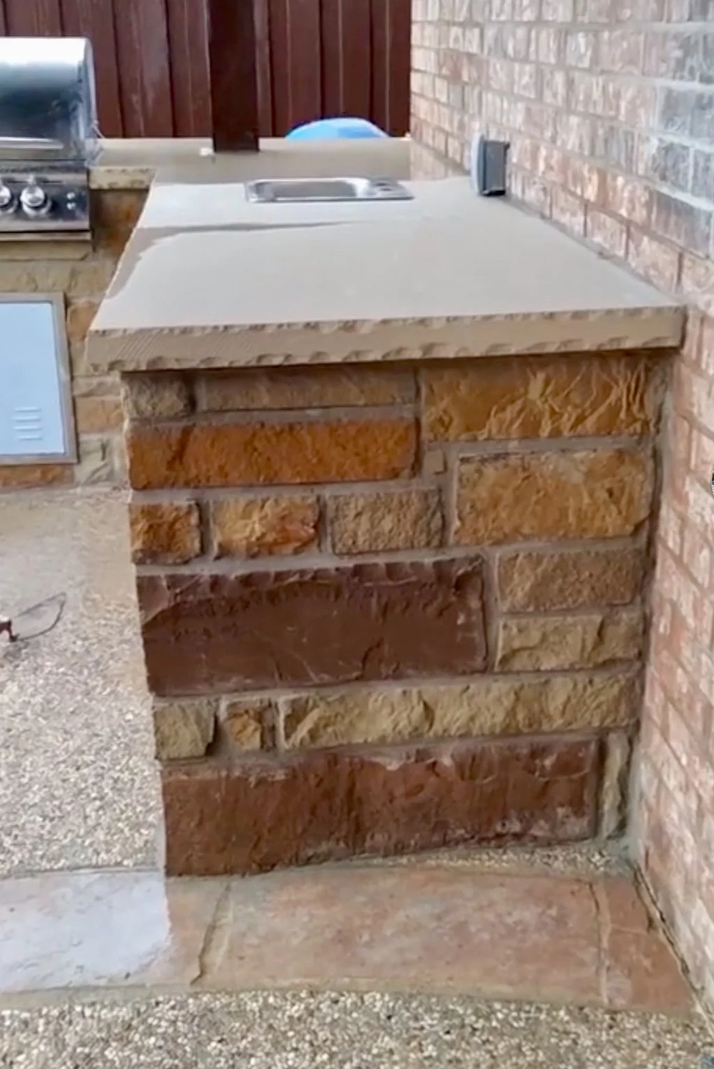 Outdoor kitchen counter with brick base, beige granite countertop, a built-in sink, and a small speaker on the wall, against a brick wall and a wooden fence.