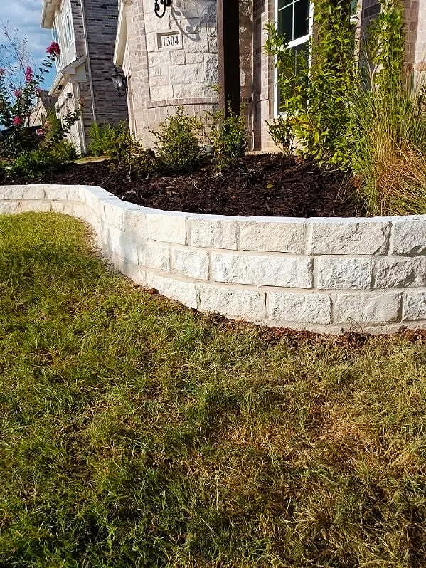 A landscaped garden bed with shrubs and plants, bordered by a white stone retaining wall in front of a house with a textured exterior wall and a black lantern-style outdoor light.