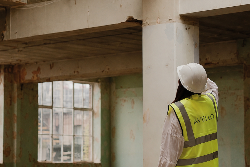 Construction worker wearing a safety vest with the word "AVELLO" and a white hard hat inspecting a room with distressed walls and a large window.