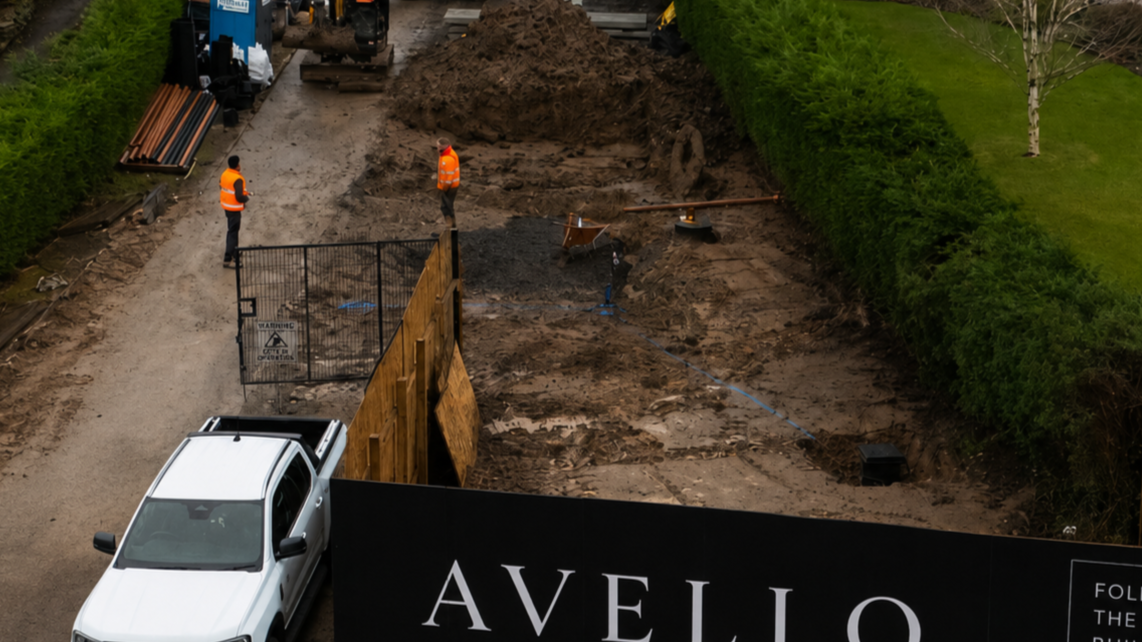 Construction site with dirt and machinery, two workers wearing orange safety vests, behind a black fence, with a white vehicle in the foreground and trees on the sides.