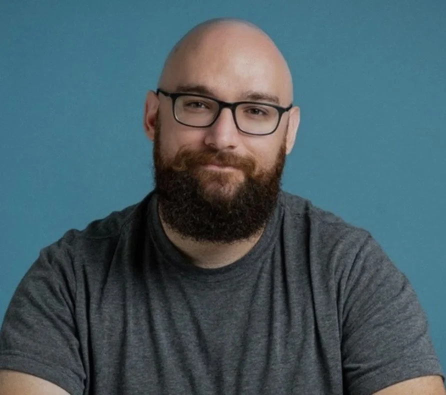 A man with glasses and a beard, wearing a gray t-shirt, sitting against a blue background.