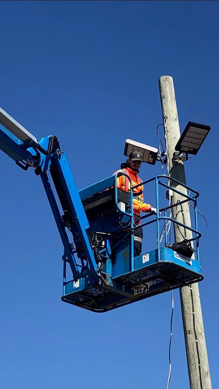 A worker in an orange safety vest and hat standing in a blue aerial platform bucket, working on a wooden utility pole with electronic equipment and solar panels attached.