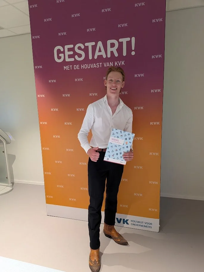 Jonge man met witte overhemd en zwarte broek, die een brochure vasthoudt, staand voor een KVK-banner met de tekst 'GESTART! MET DE HOUVAST VAN KVK'.